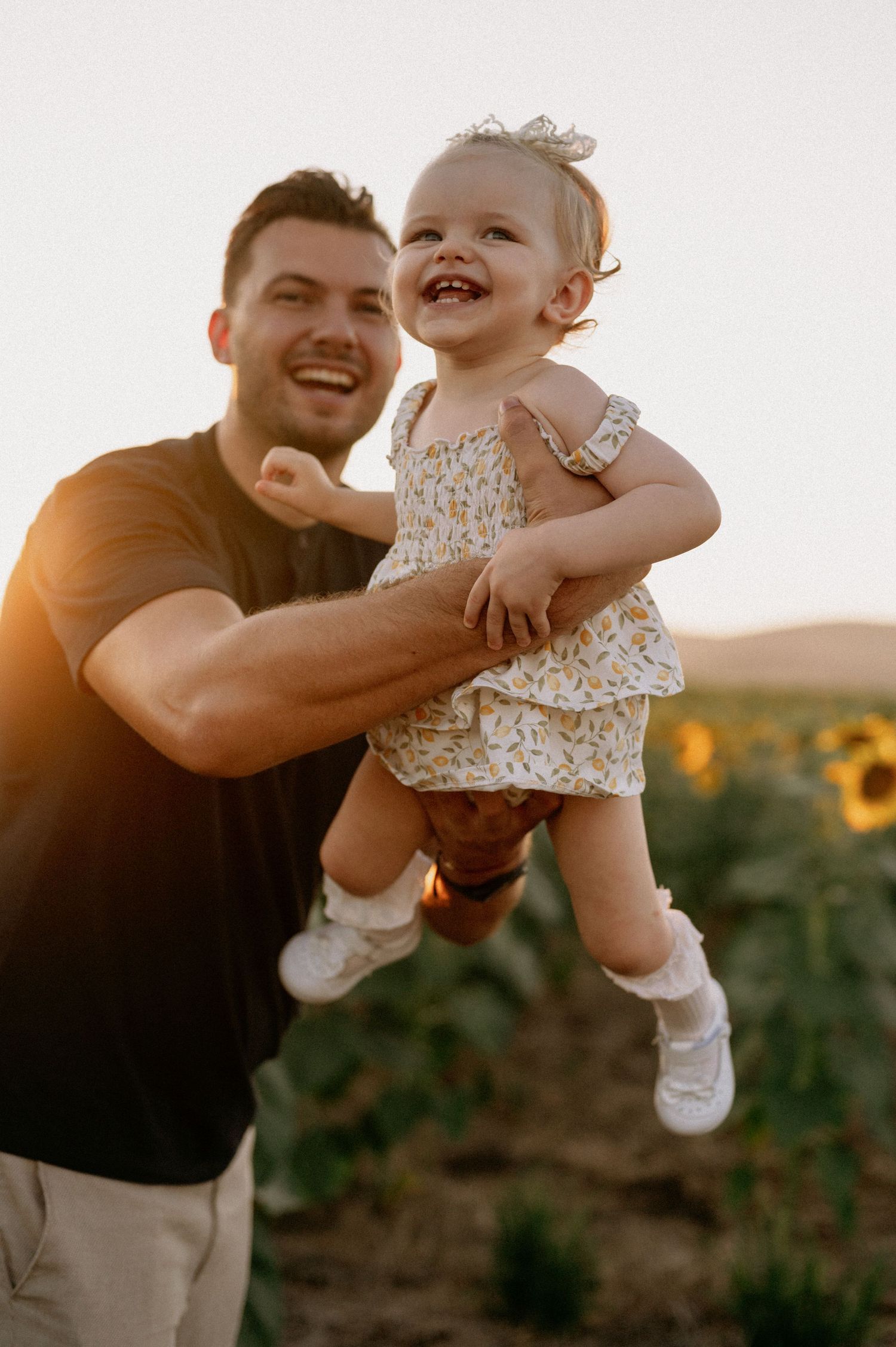 A joyful father lifts his laughing baby girl in a sunflower field during golden hour sunset.