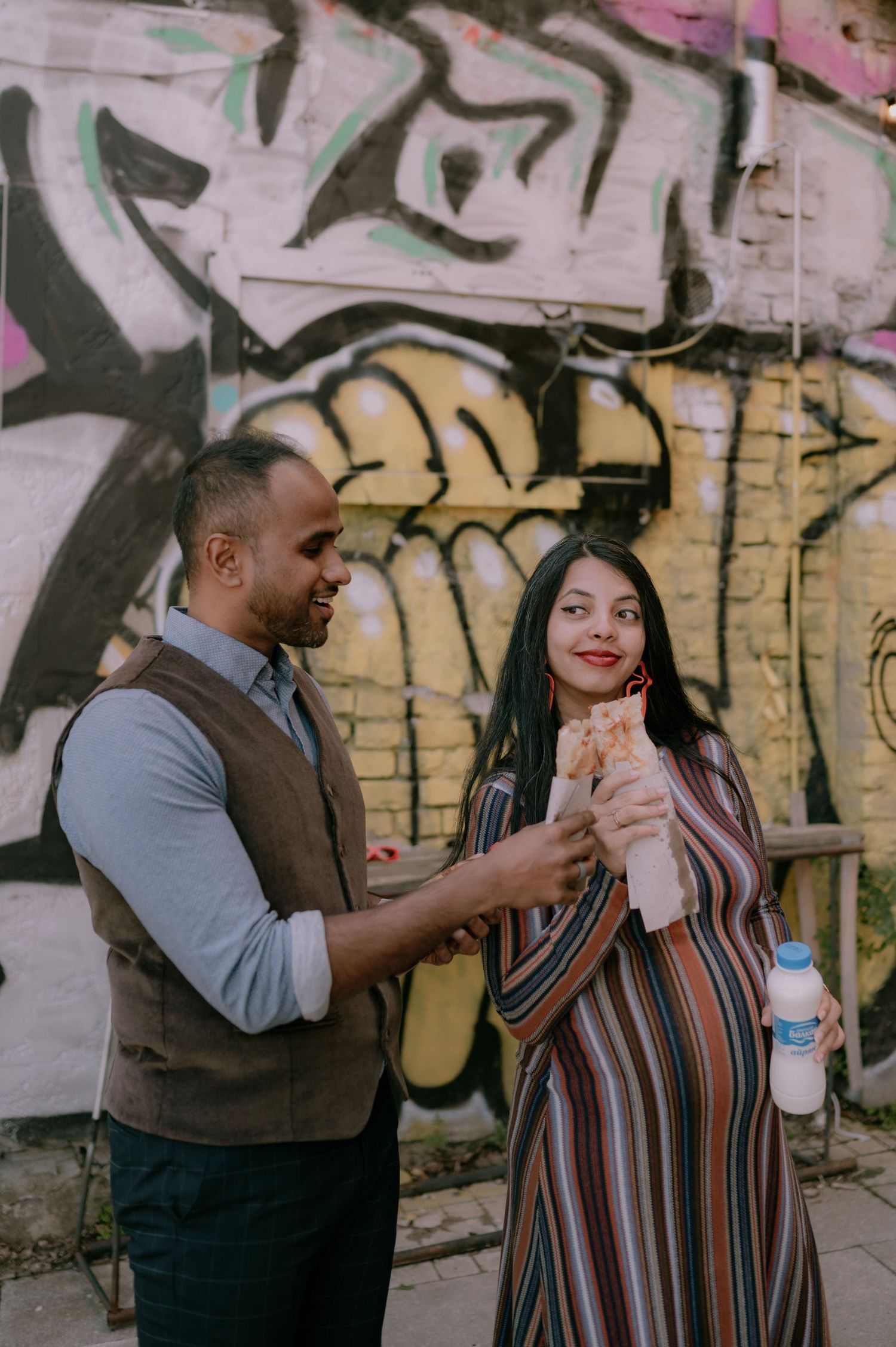 A man and pregnant woman share food in front of a colorful graffiti wall outdoors.