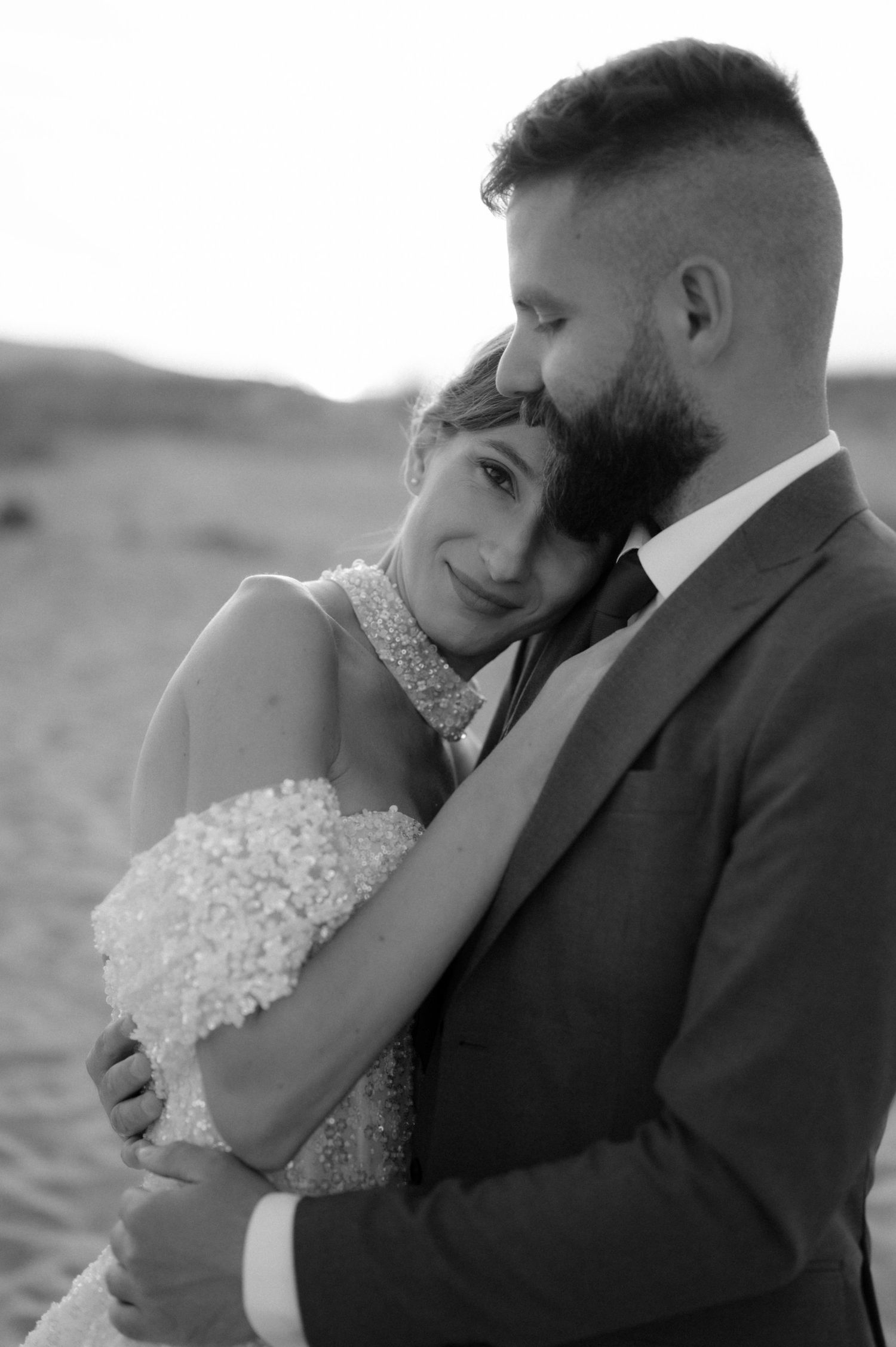 Black and white wedding photo of a couple embracing, bride in beaded gown leaning on groom in suit outdoors.