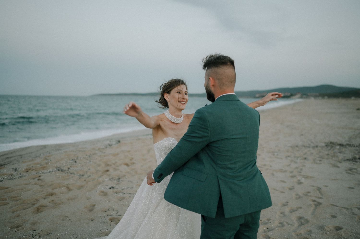 Bride and groom dancing joyfully on a sandy beach with ocean waves in the background.