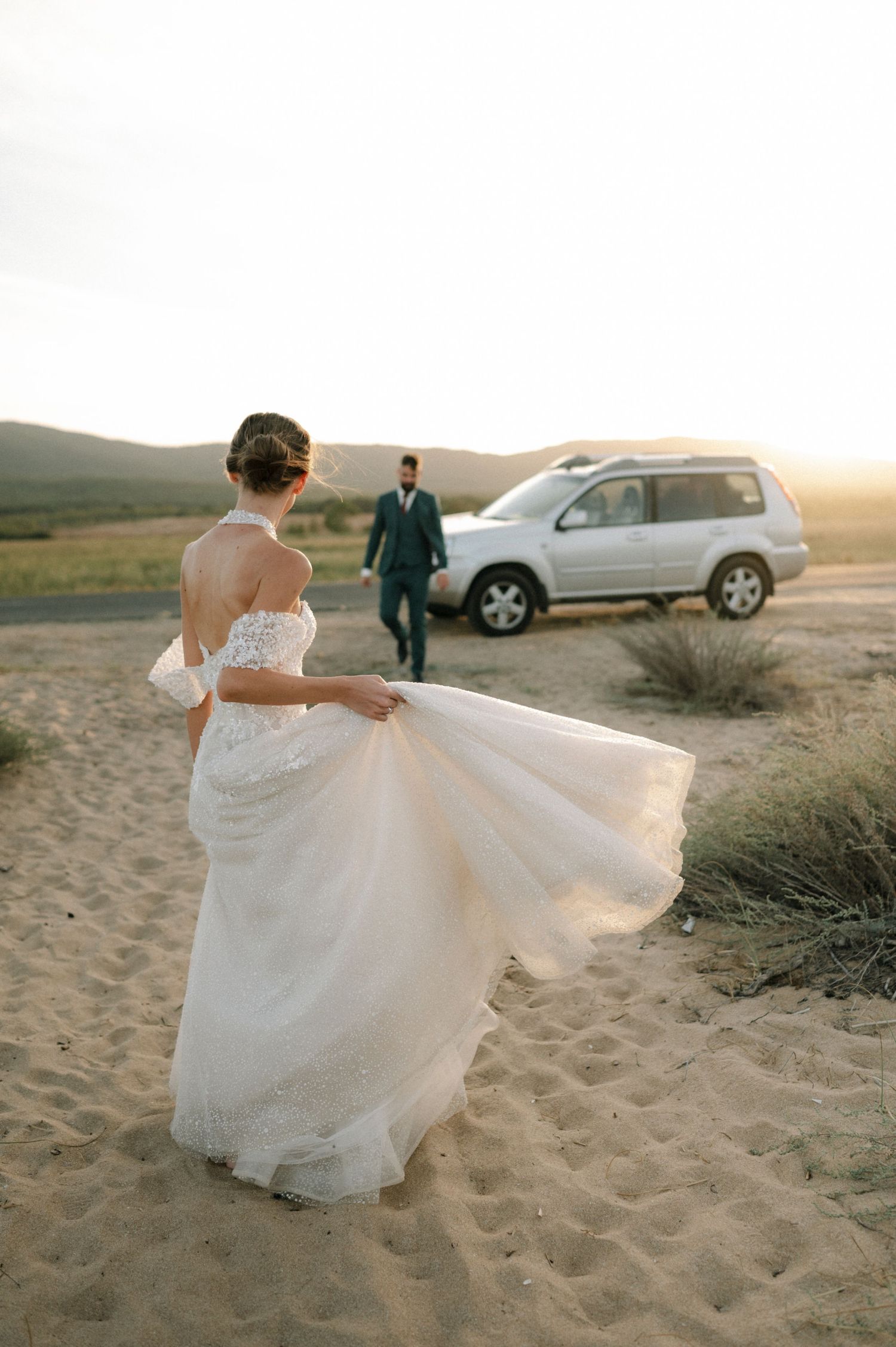 Bride in white gown holds dress on sandy desert at sunset, groom in teal suit walks from SUV.