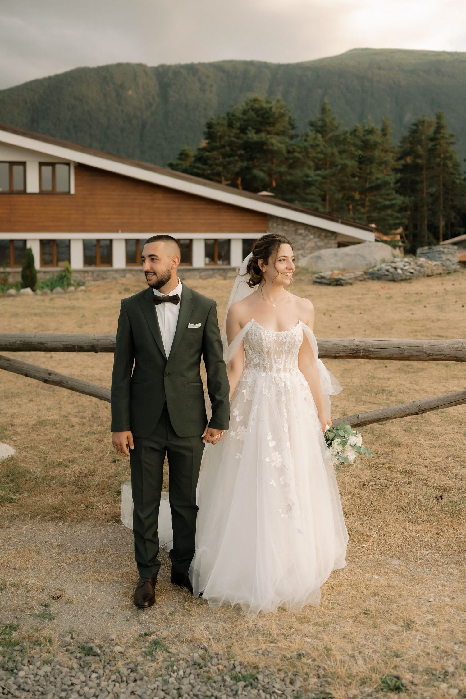 A couple walks hand in hand across a scenic mountain landscape during their wedding photoshoot.