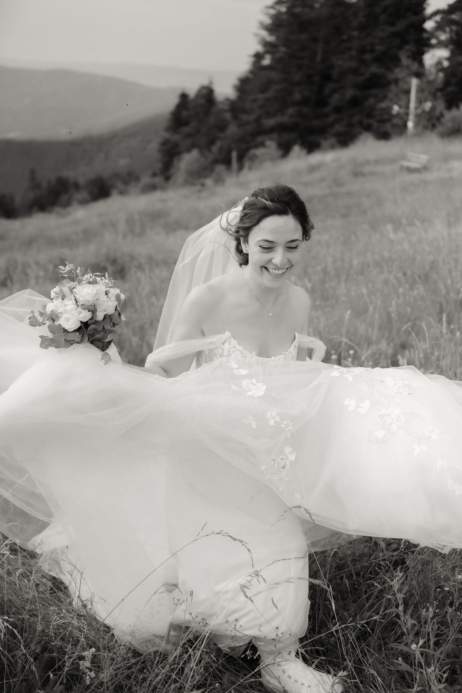 A bride in a flowing white dress lies gracefully in a grassy meadow with mountains in the background.
