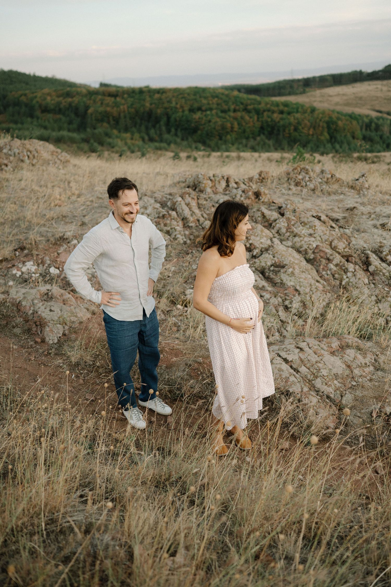A couple stands together in a grassy field with rolling hills and forest in the background during golden hour.