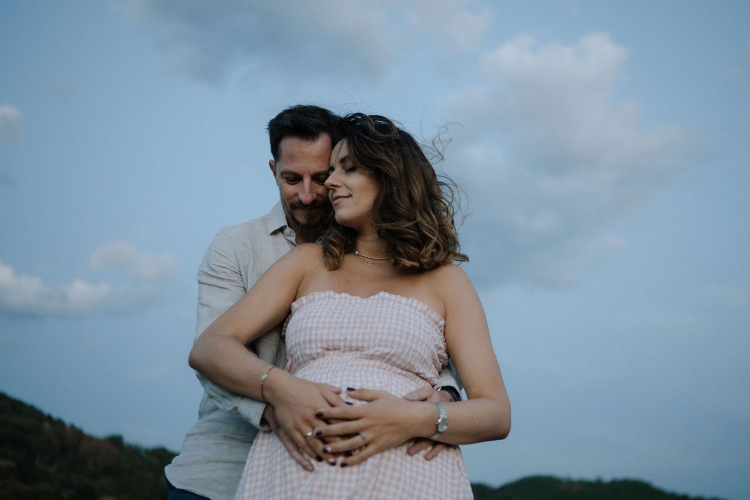 A couple embraces during a maternity photoshoot against a cloudy blue sky and mountain backdrop.