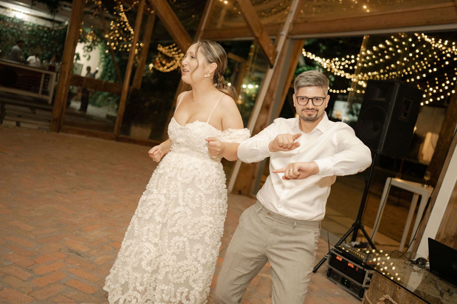 A couple dances together at an evening wedding reception under twinkling string lights in a rustic venue.