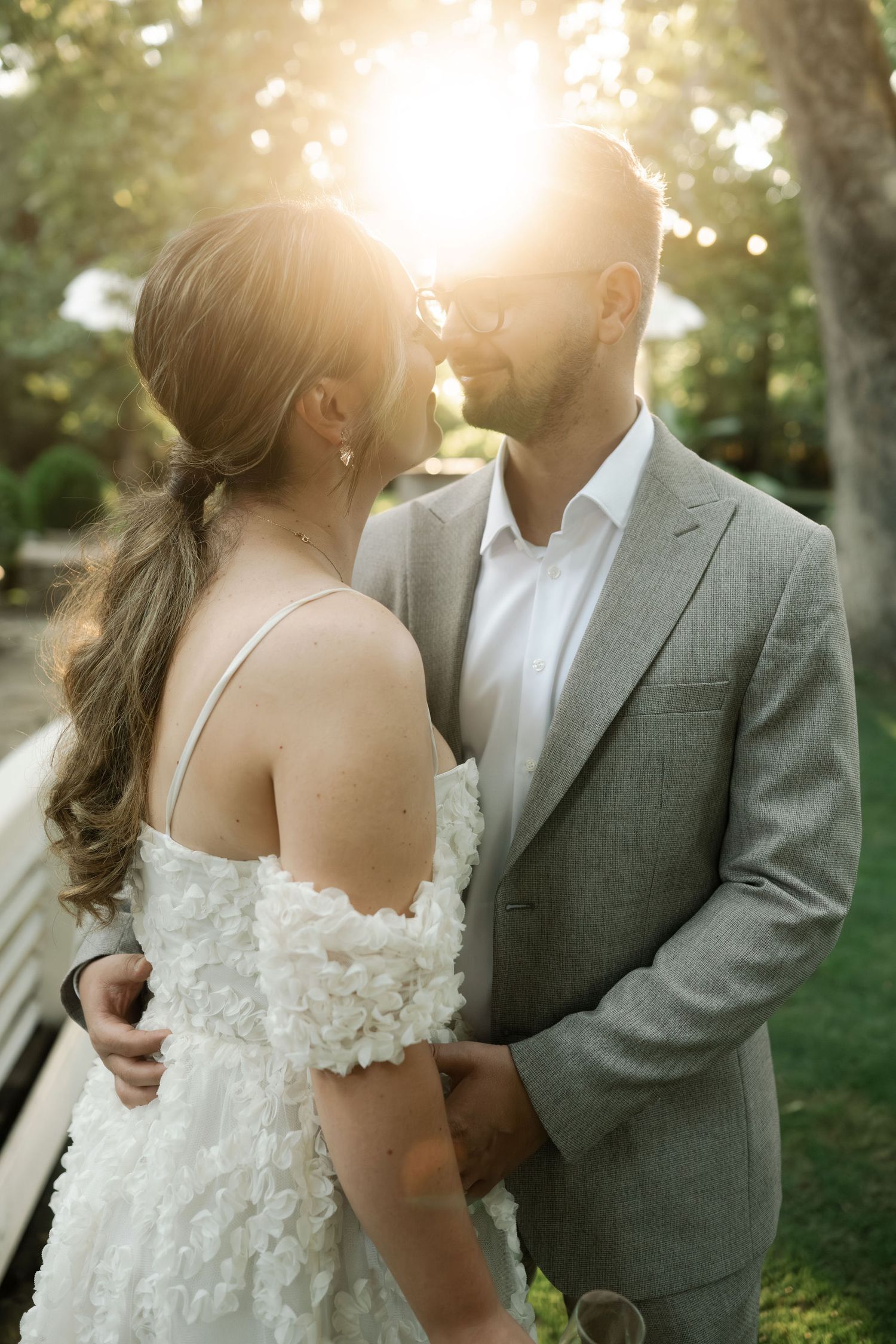 A couple in wedding attire share an intimate moment in a sunlit garden setting with trees in the background.
