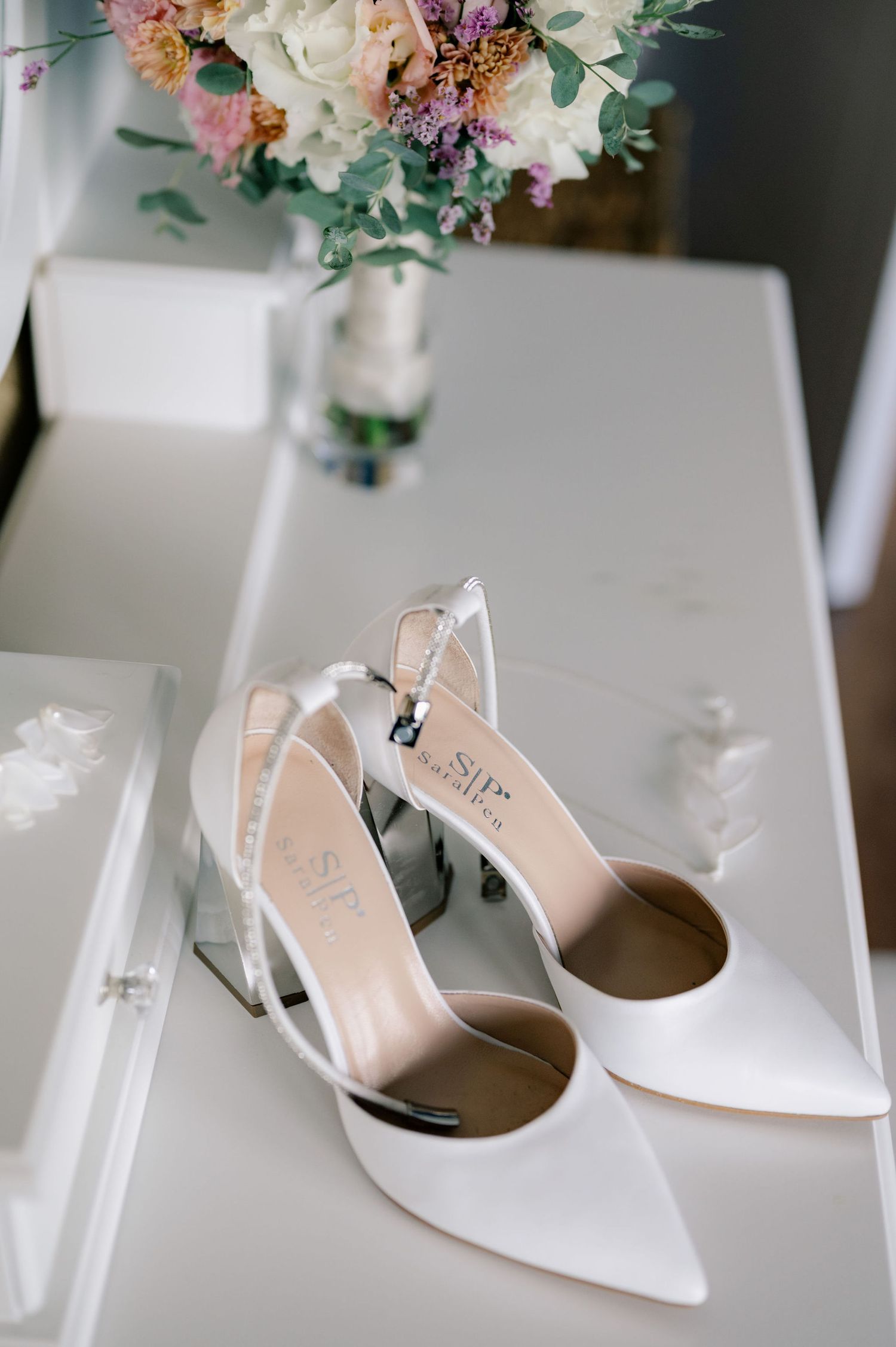 Elegant white bridal shoes with pointed toes and clear block heels displayed next to a delicate floral bouquet.