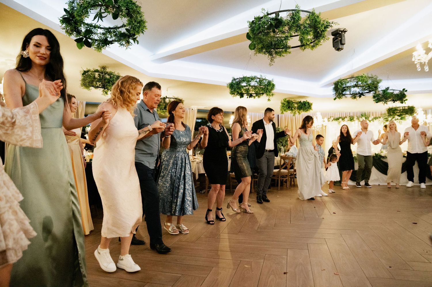 Wedding guests dance in a line during a reception in a white tent venue with hanging greenery decorations.