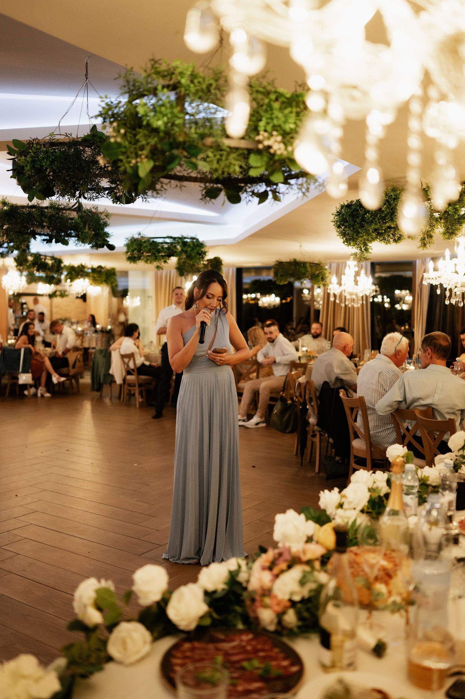 An elegant wedding reception venue with hanging greenery, chandeliers, and floral centerpieces on tables.