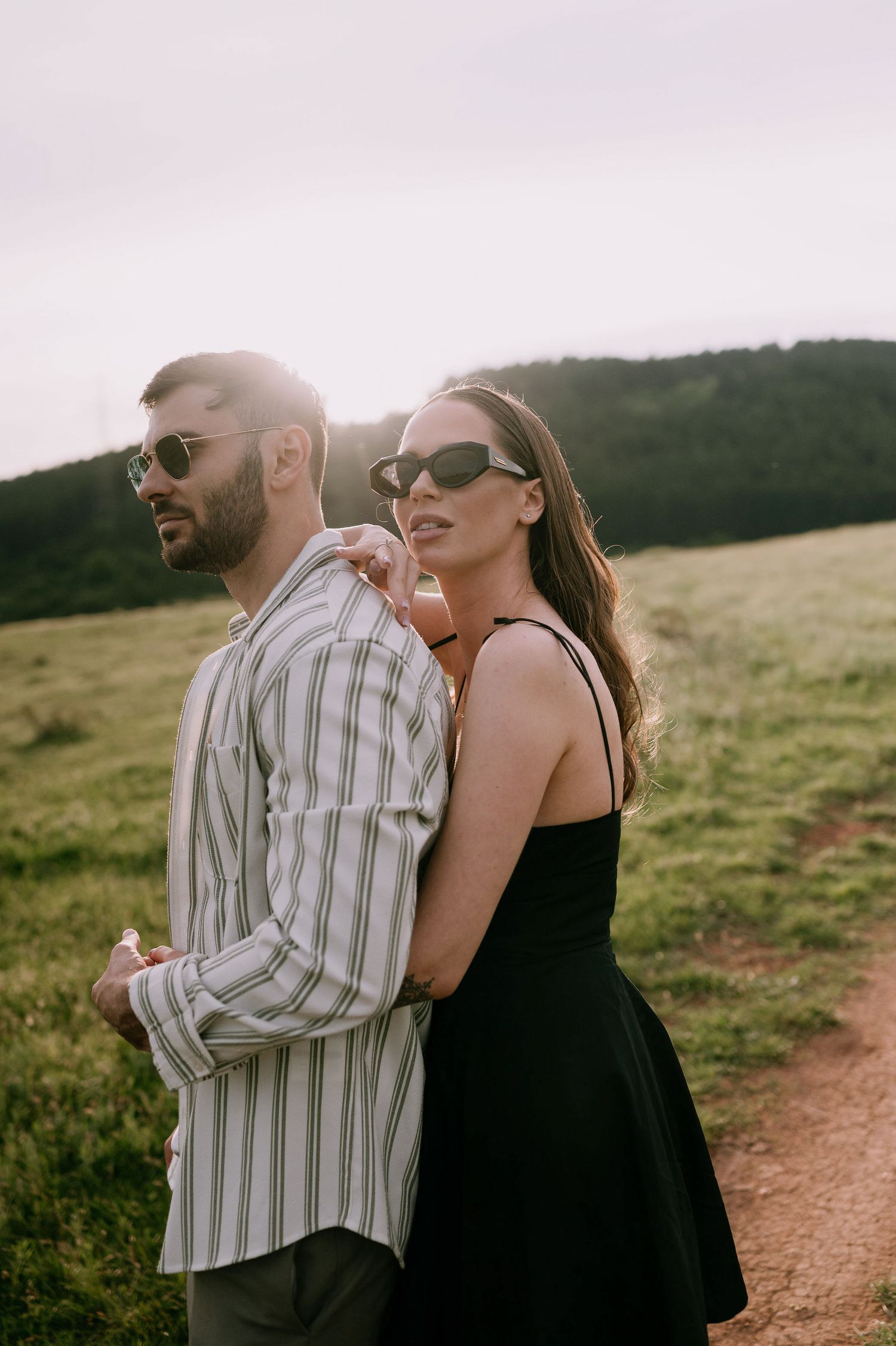 A couple laughs together while walking along a dirt path through a grassy hillside on a sunny afternoon.