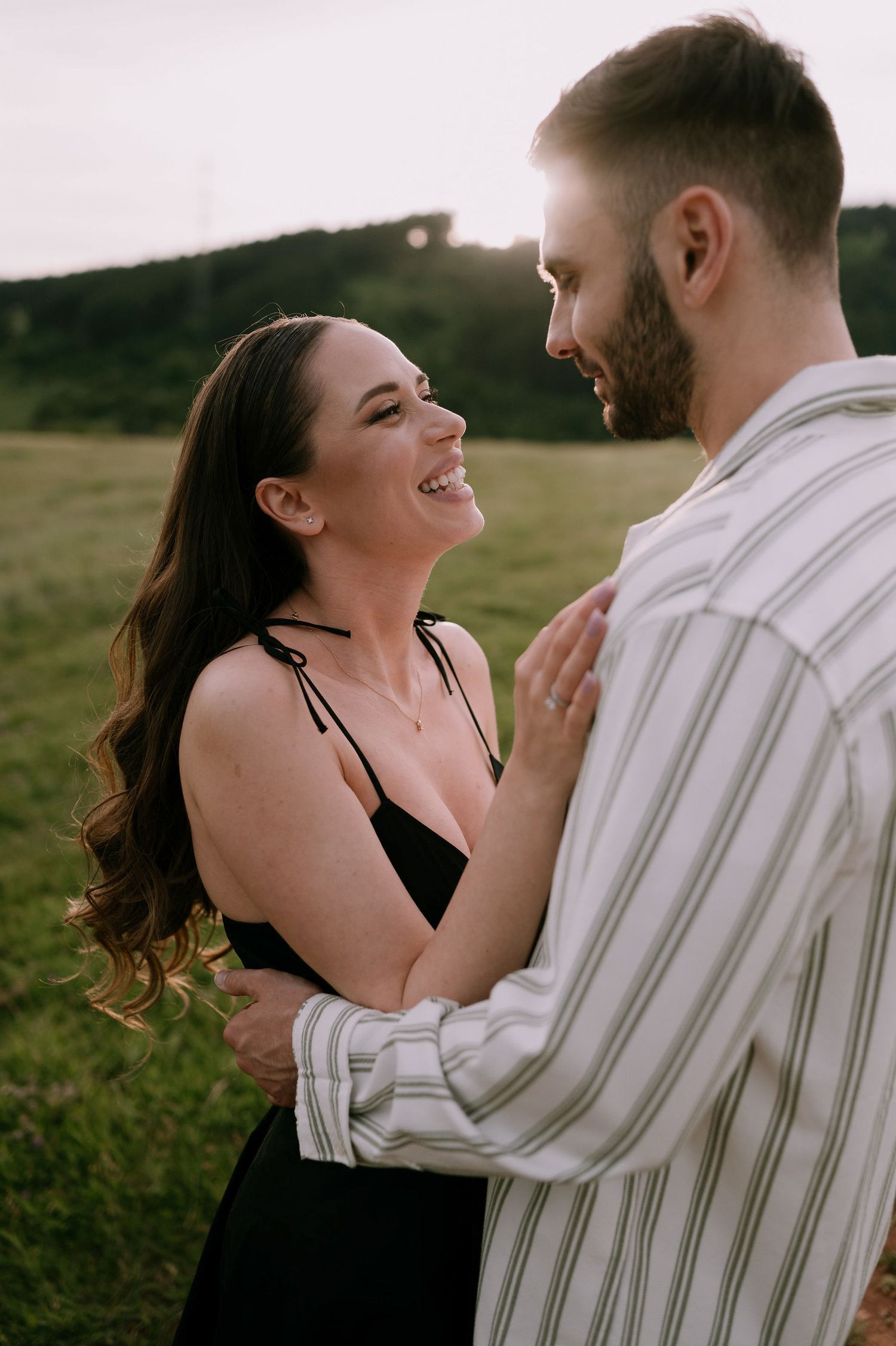A romantic couple embraces and laughs together during a sunset photoshoot in an open field.
