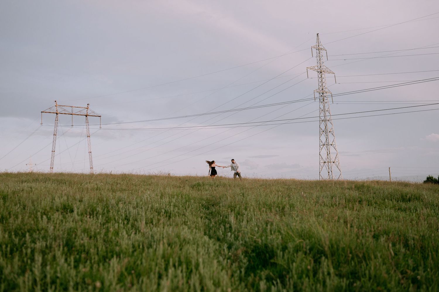 Two silhouettes walk along a grassy hilltop with power transmission towers against a cloudy sky.