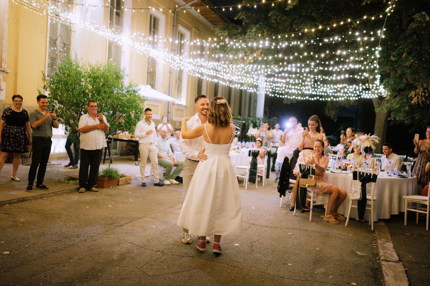 An outdoor evening wedding reception with string lights overhead as a couple dances on a stone patio near dining tables.