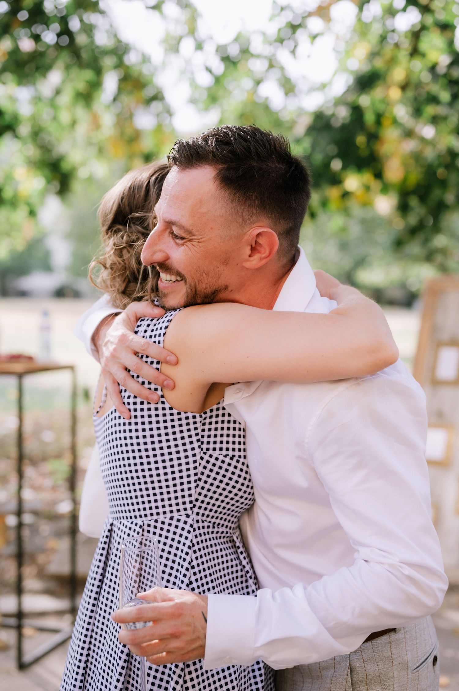 Two people share a warm embrace outdoors with sunlit trees and foliage in the background.