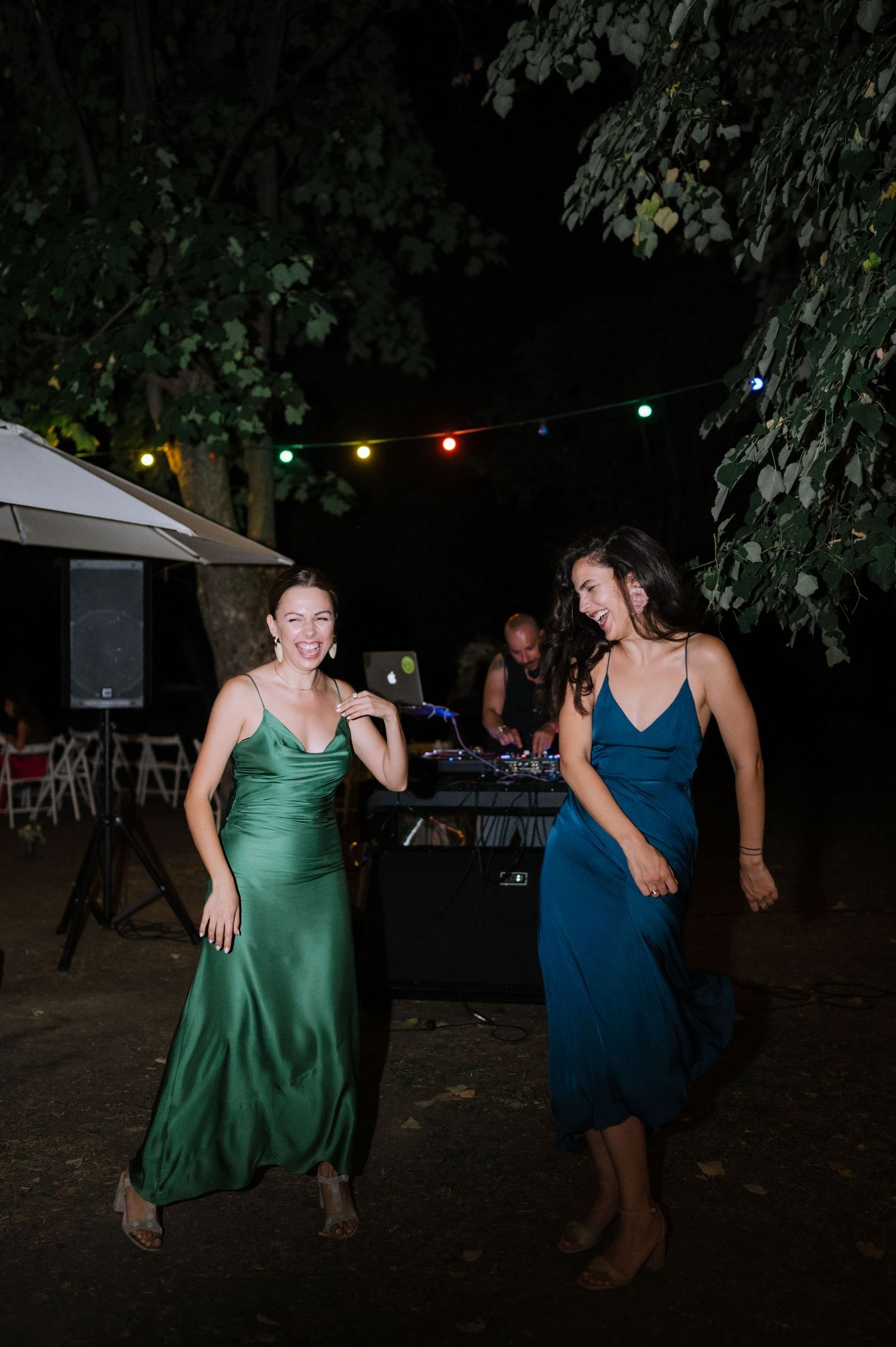 Two people in formal dresses dance joyfully at an outdoor nighttime party with string lights visible in background.