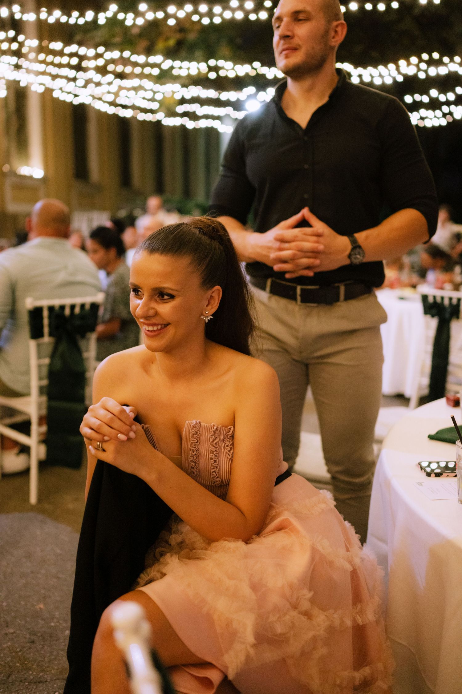 A person in a pink tulle dress laughs joyfully at a wedding reception under twinkling string lights.