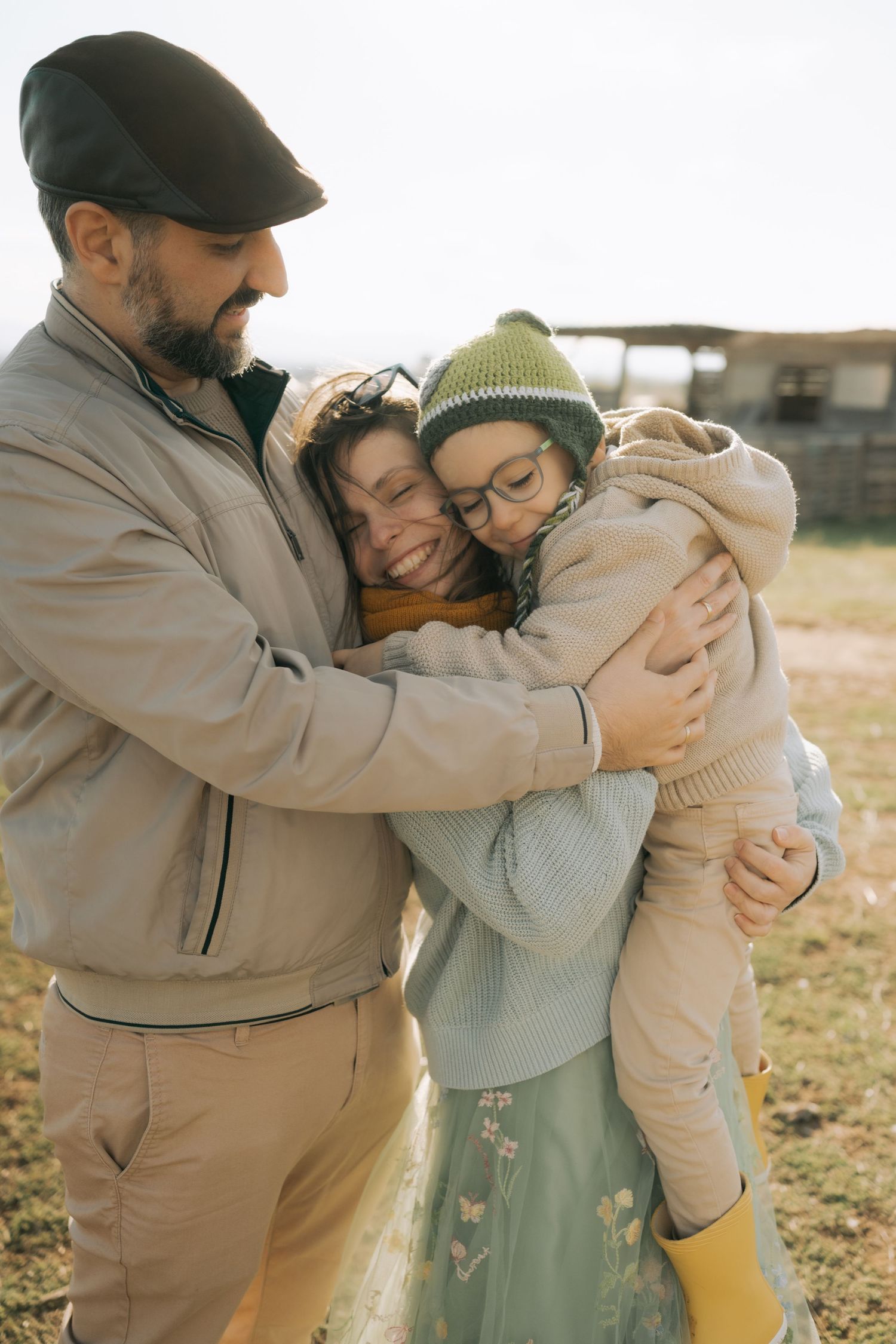 A family shares a warm embrace outdoors on a sunny day, wearing cozy winter attire and showing affection.