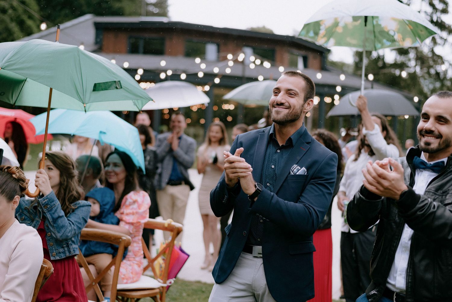 Wedding guests hold turquoise umbrellas during an outdoor ceremony on a rainy day.
