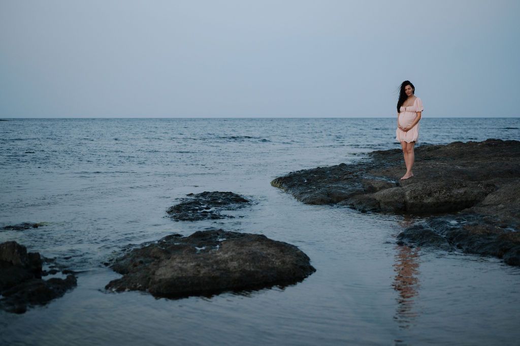 Person standing on rocky coastal outcrop at sunset with waves crashing against dark volcanic rocks near ocean shore.