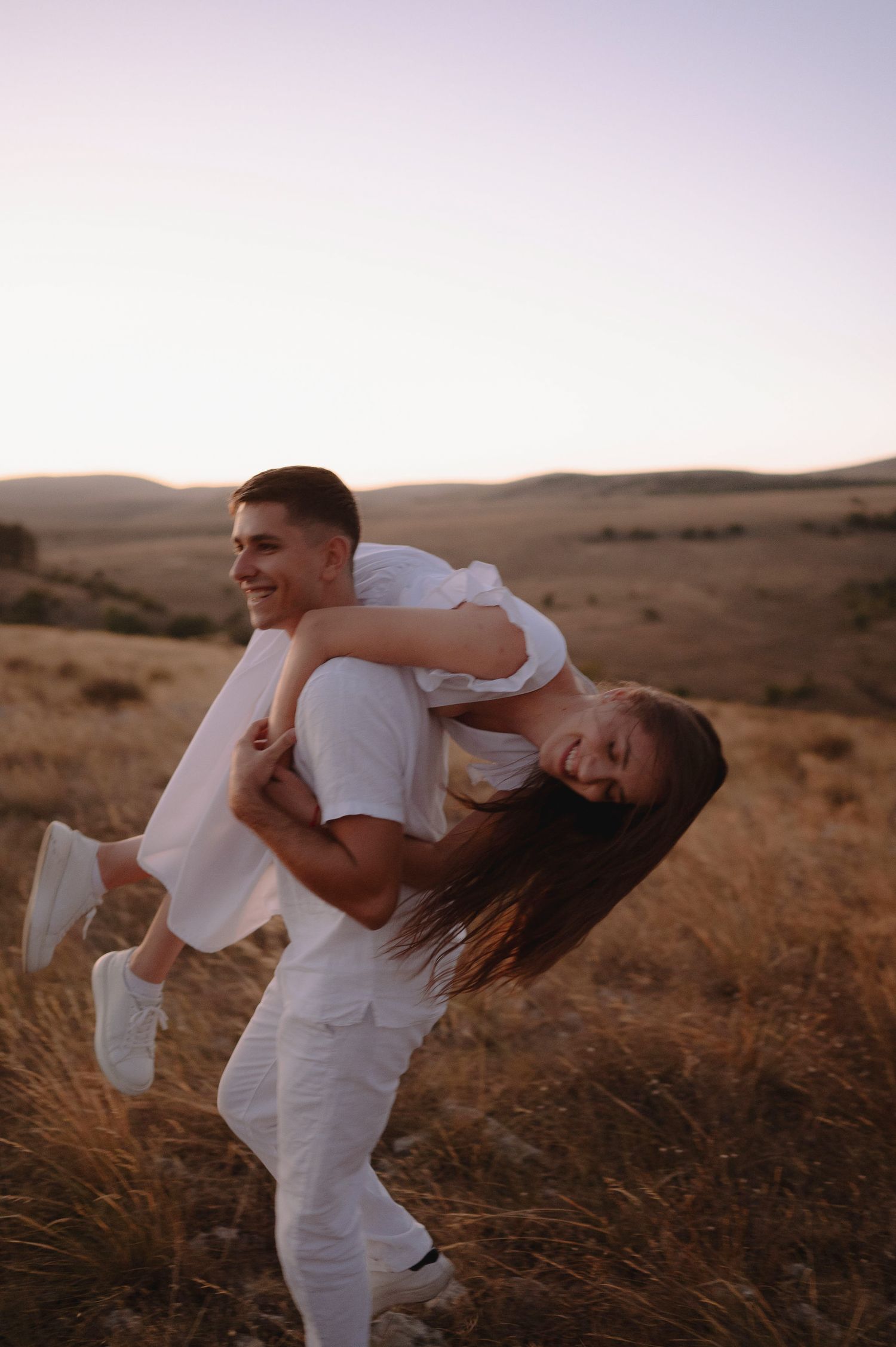 A couple in white outfits playfully poses together on a hillside during golden hour.