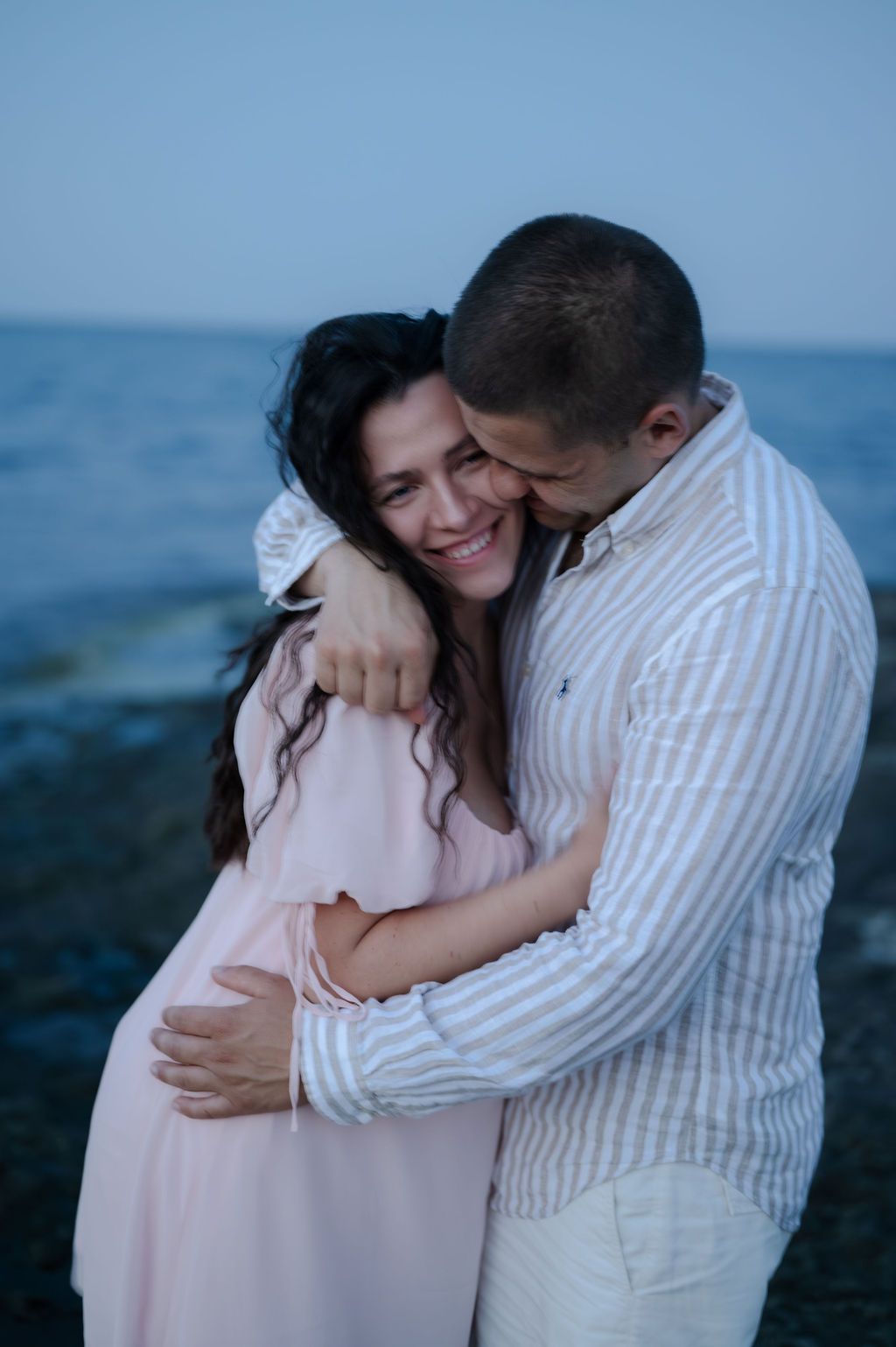 A couple shares tender moments at the beach during sunset, wearing pink and white outfits while embracing each other.