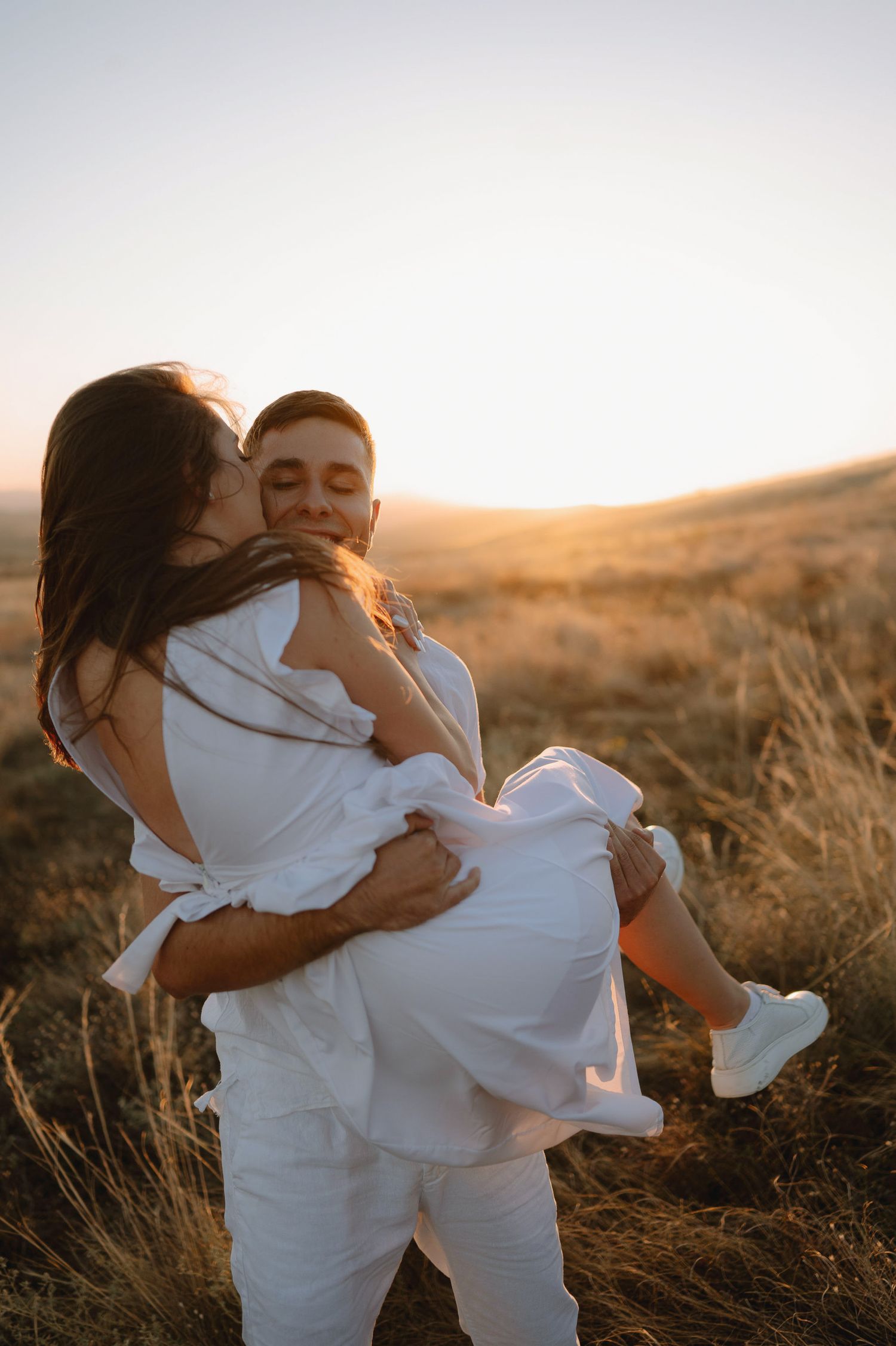 Romantic moment captured between a couple in white outfits against a sunset-lit hillside backdrop.