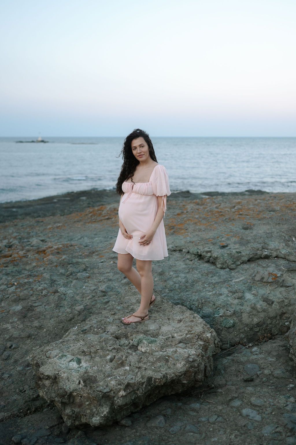 A person in a pink dress stands on coastal rocks at dusk with the ocean in the background.