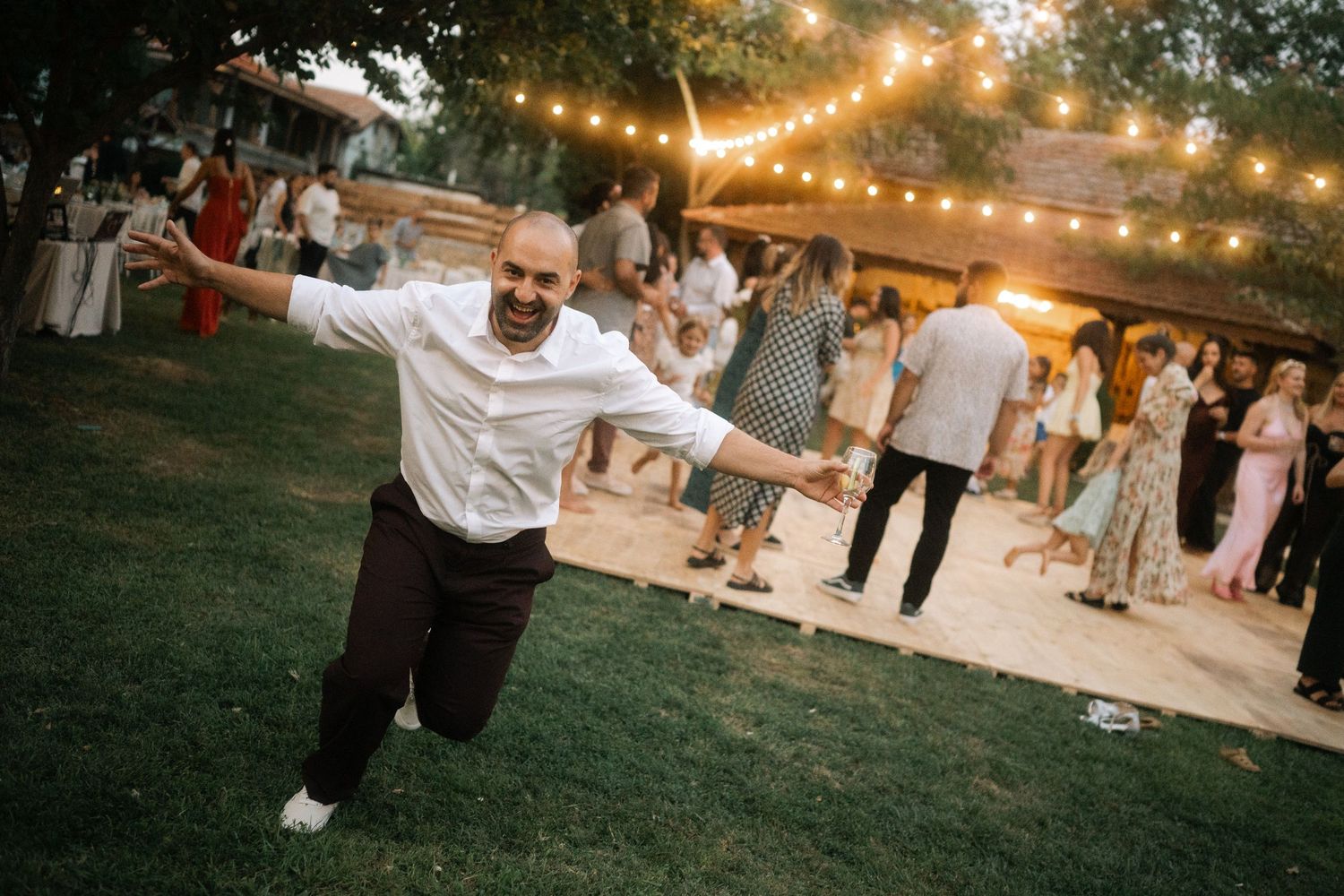 A person dances joyfully on a lawn at an outdoor evening party with string lights glowing overhead.