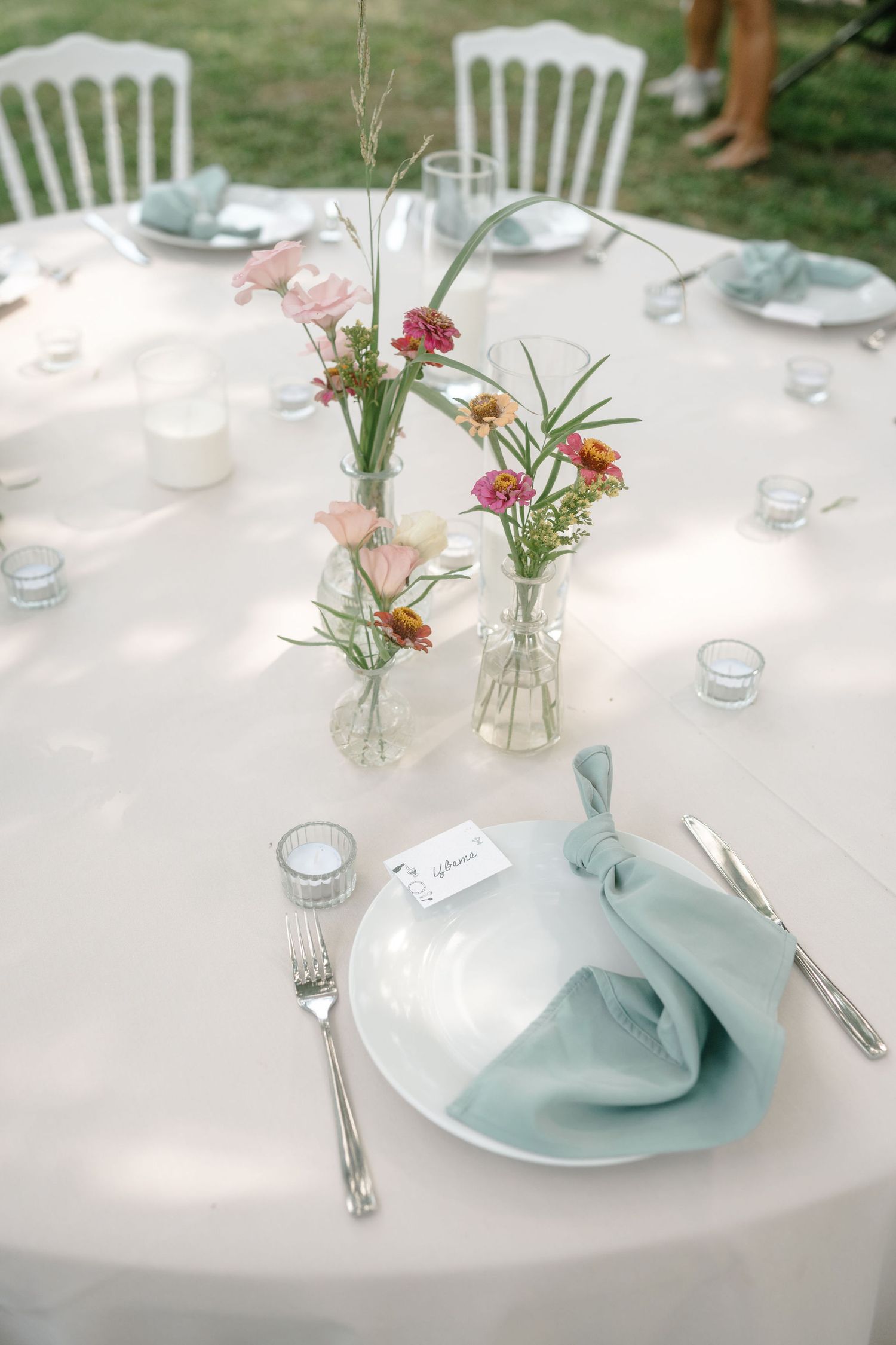 Elegant wedding table settings with soft blue napkins and delicate floral centerpieces in small glass vases on white tablecloths.