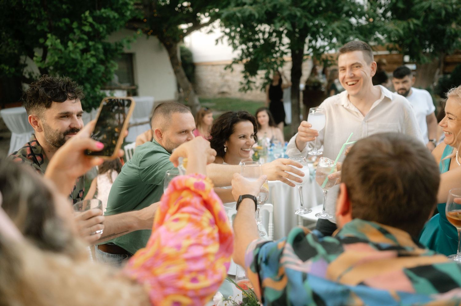 Wedding guests seated at outdoor tables celebrate during a summer reception under trees and string lights.