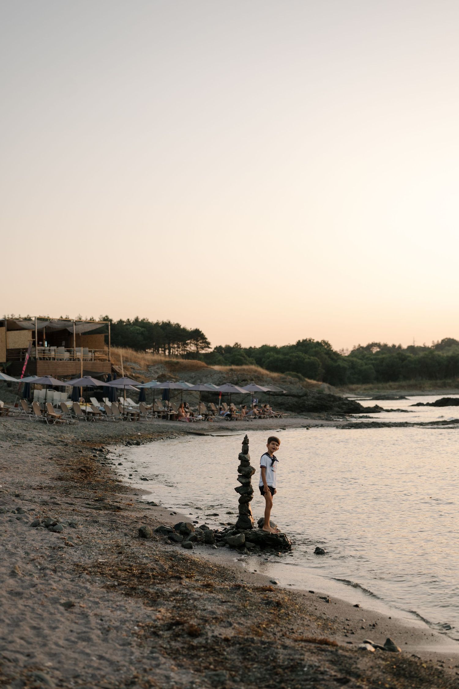 Two silhouettes stand on a rocky beach at sunset with calm waters and coastal buildings in the background.