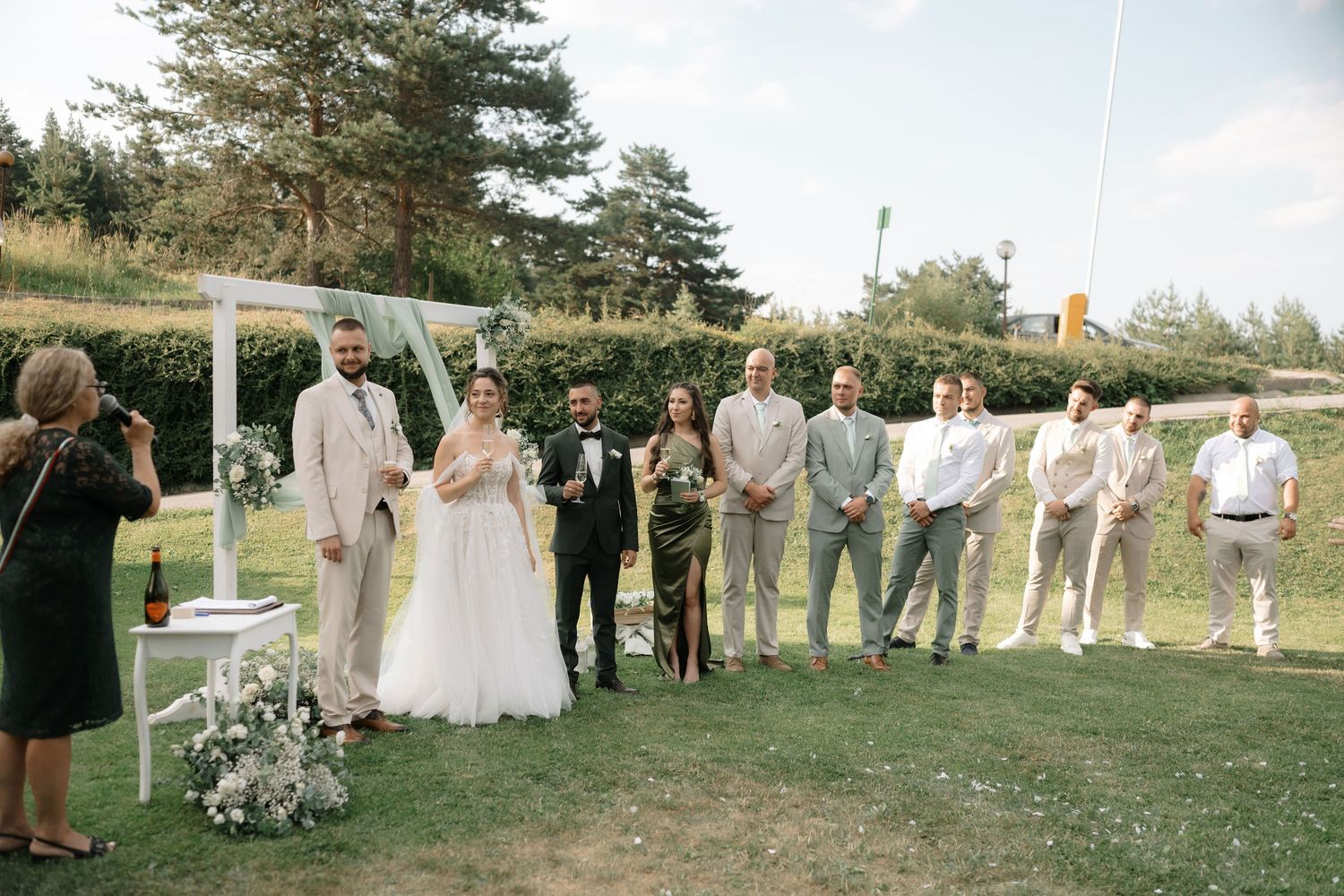 Wedding party gathers outdoors on a grassy lawn with white floral arch and natural decor during daytime ceremony.