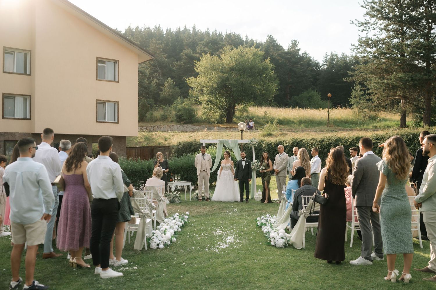 Wide angle shot of wedding attendees standing in rows on a lawn with trees and buildings in the background.