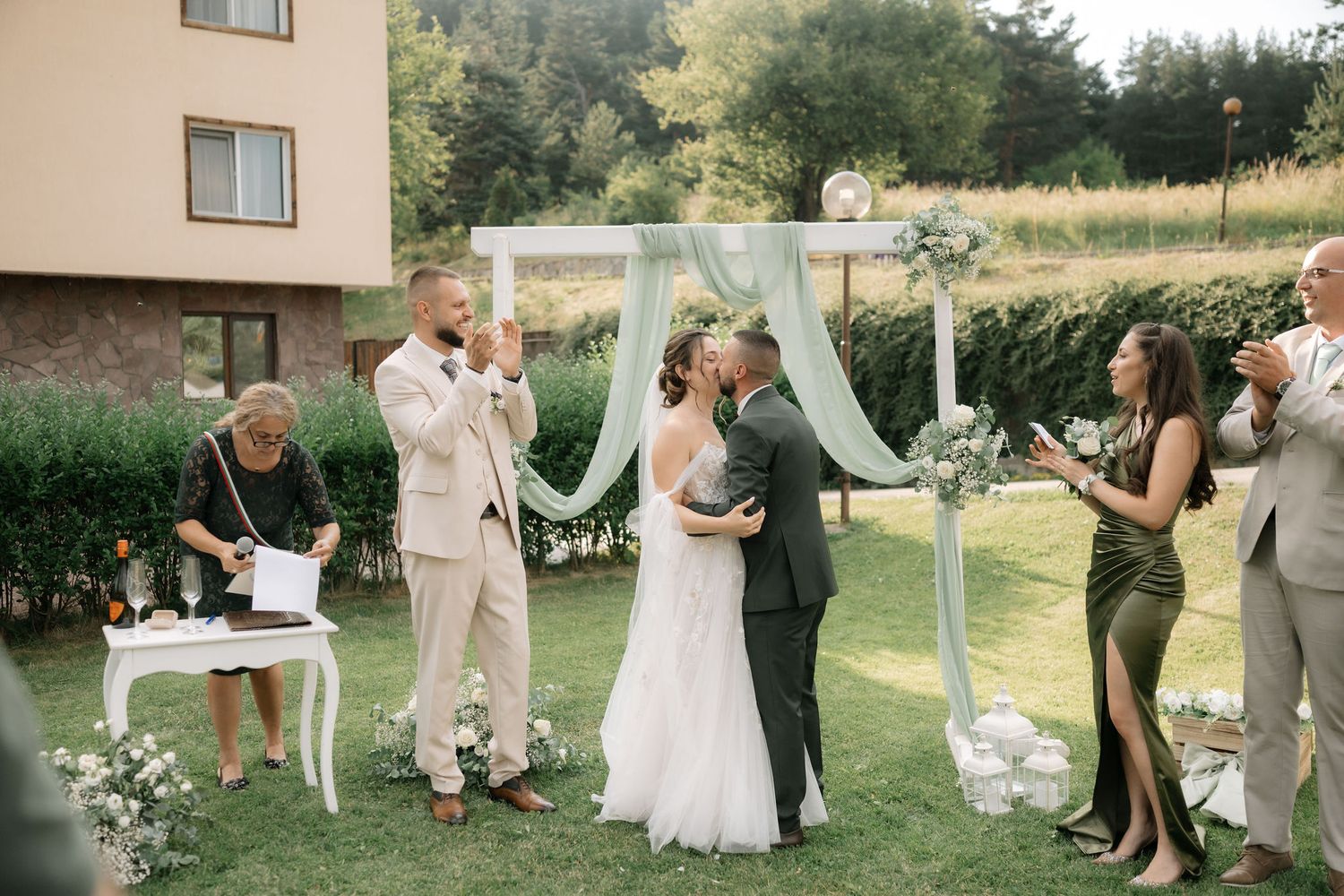 An intimate outdoor wedding ceremony takes place under a light blue fabric arch with guests celebrating on a sunny day.