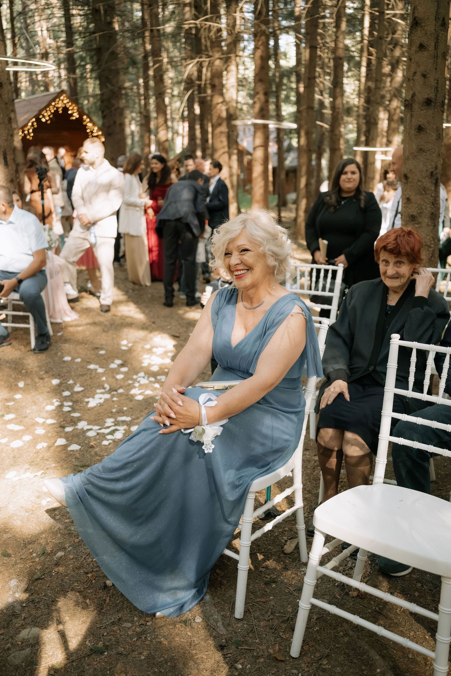 Elegant older woman in blue gown smiling at outdoor forest wedding ceremony with white chairs and guests.