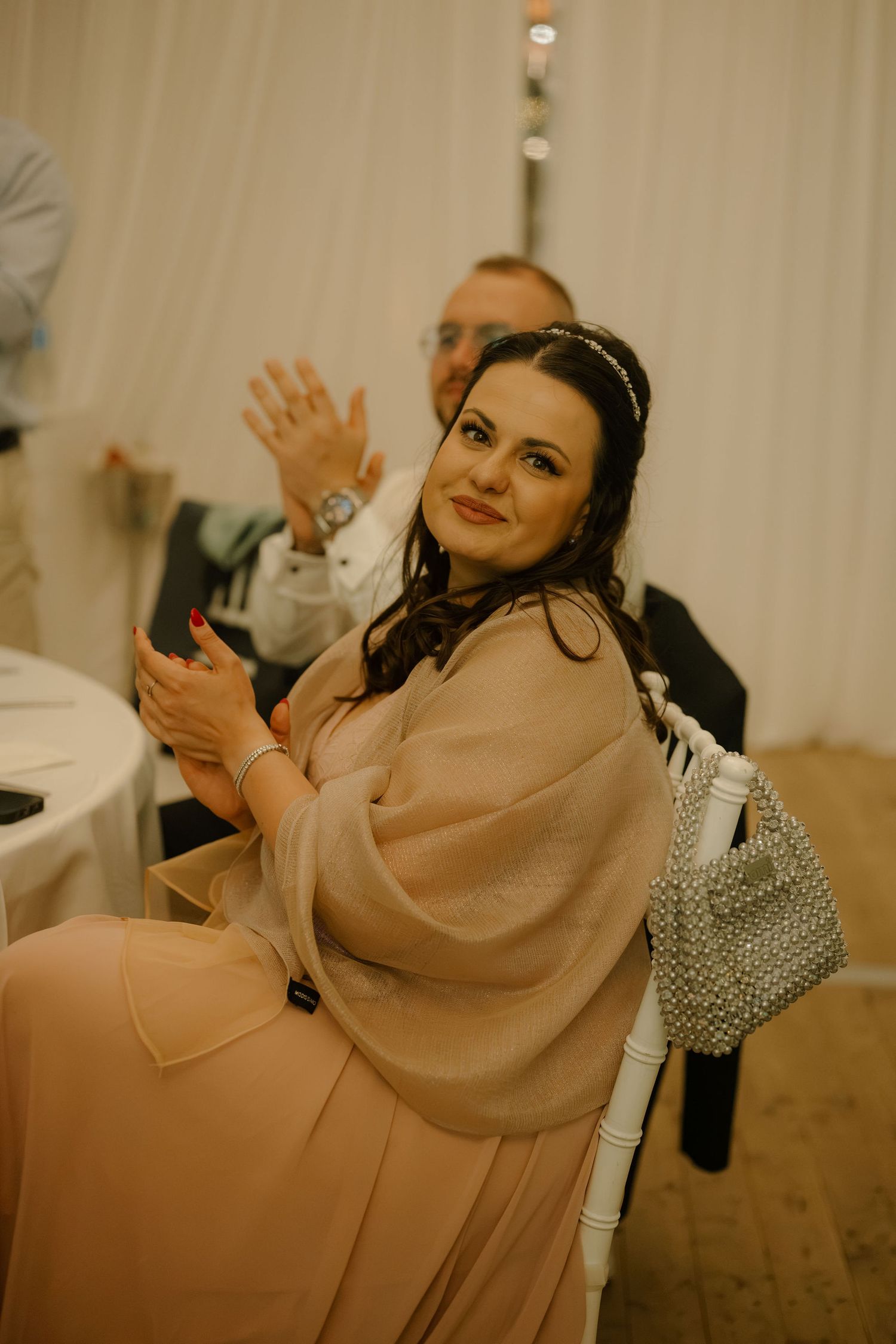 A smiling woman in a beige gown with a shawl waves at a wedding reception, seated on a white chair.