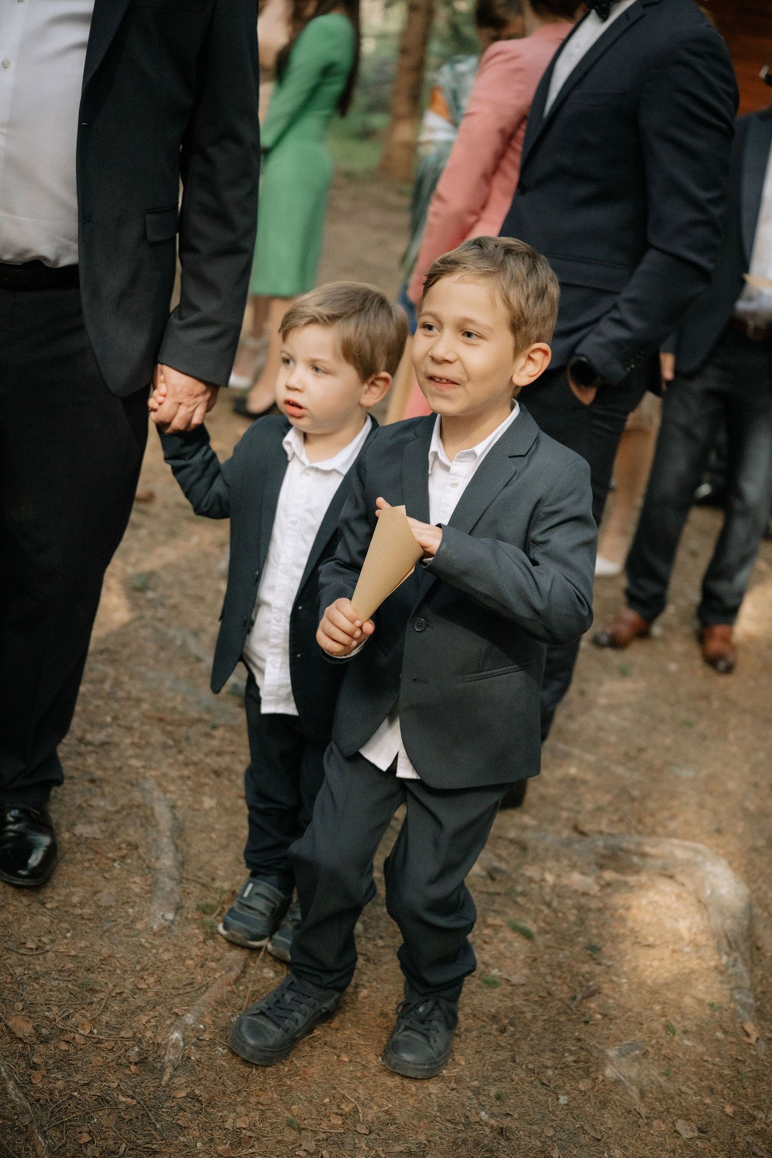 Two young boys in dark suits holding hands at an outdoor wedding ceremony, smiling and holding paper cones.