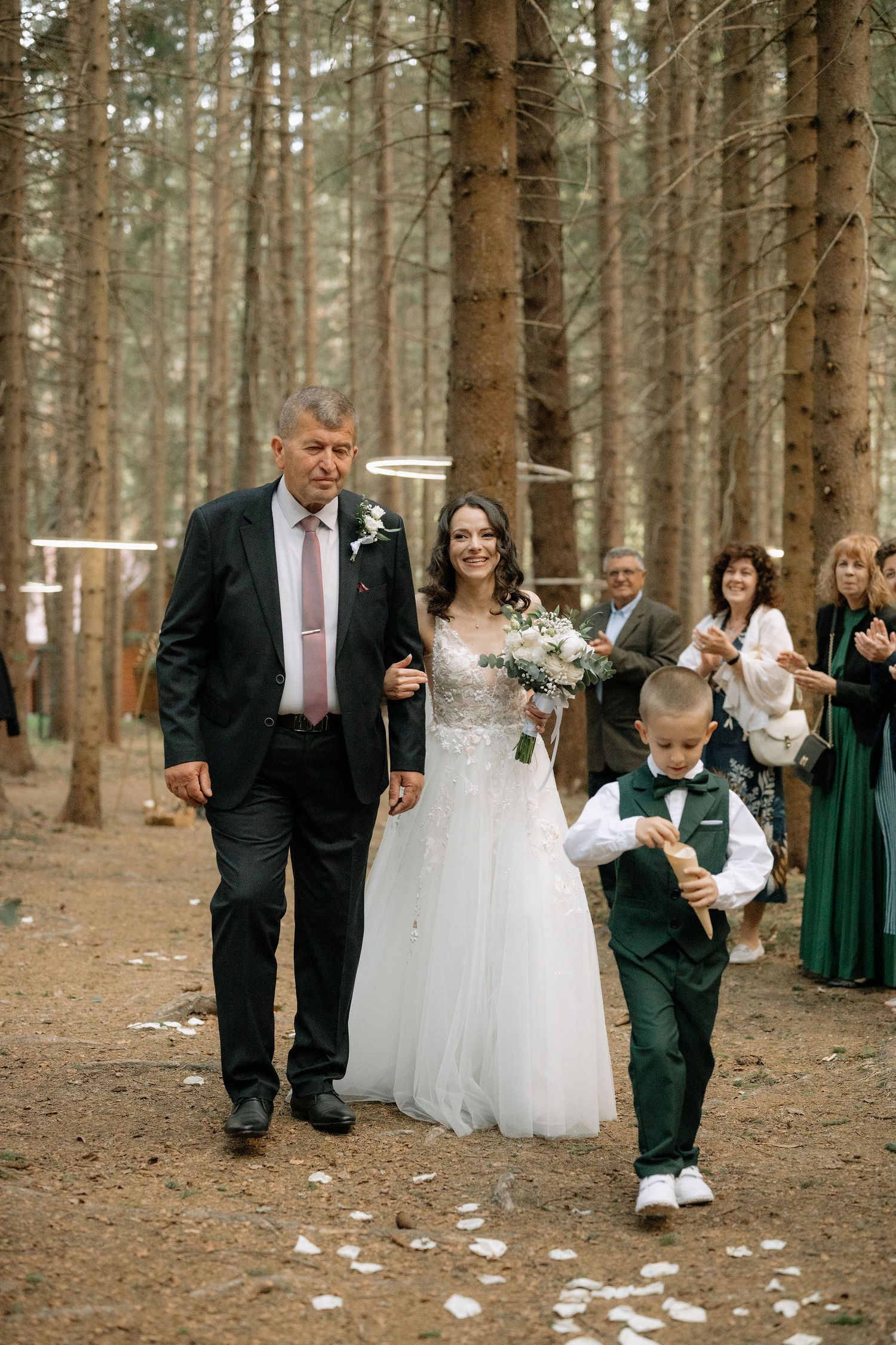 Bride walks down forest aisle with father and ring bearer during outdoor woodland wedding ceremony.