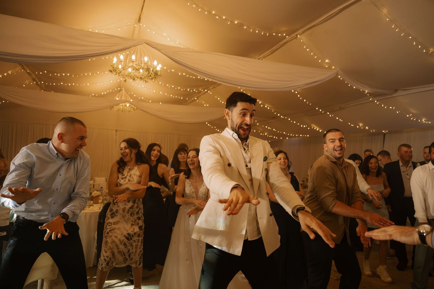 Guests dancing energetically at a wedding reception under warm fairy lights and draped ceiling decor.