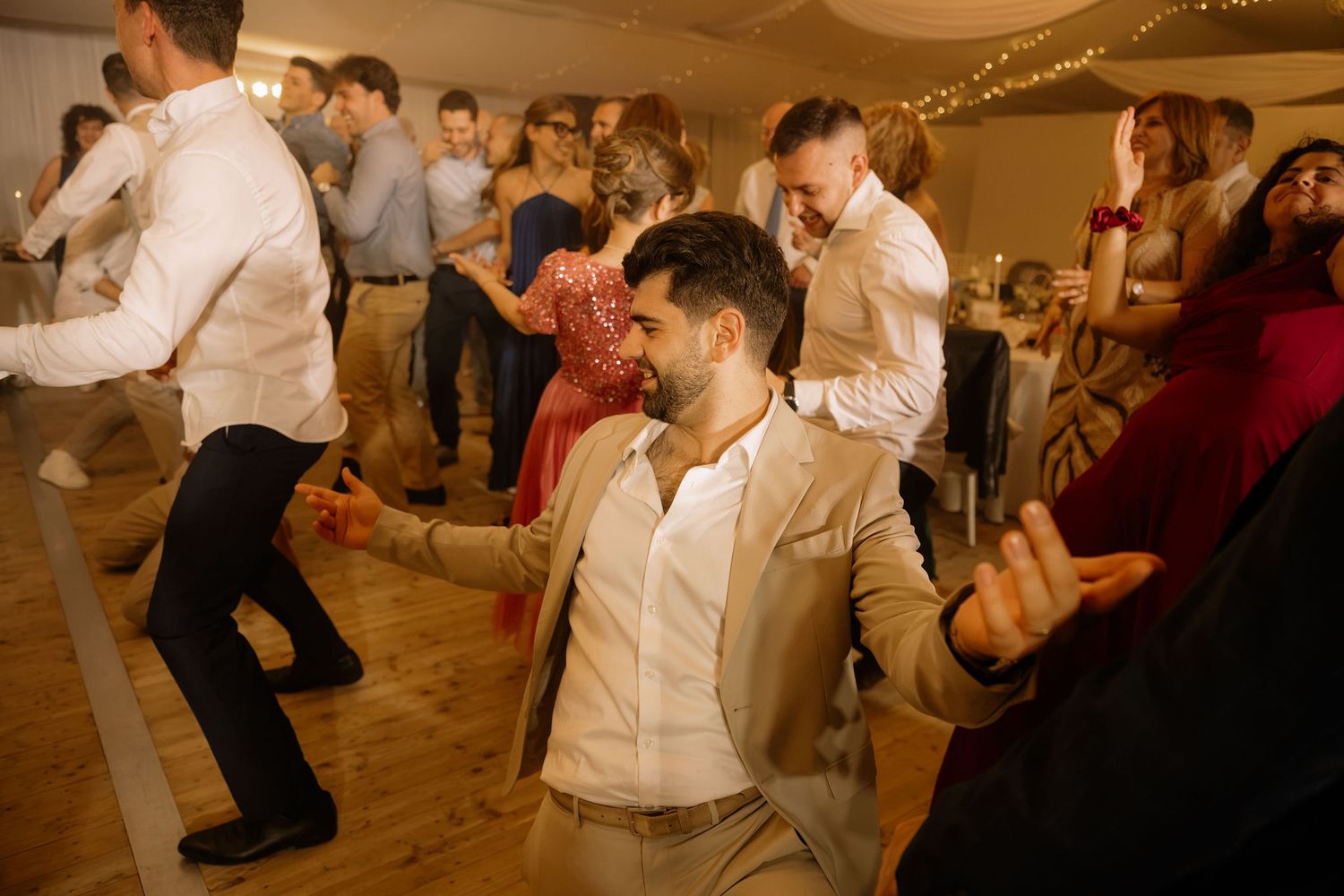 Guests dancing joyfully at a wedding reception, with a man in a beige suit smiling in the foreground.