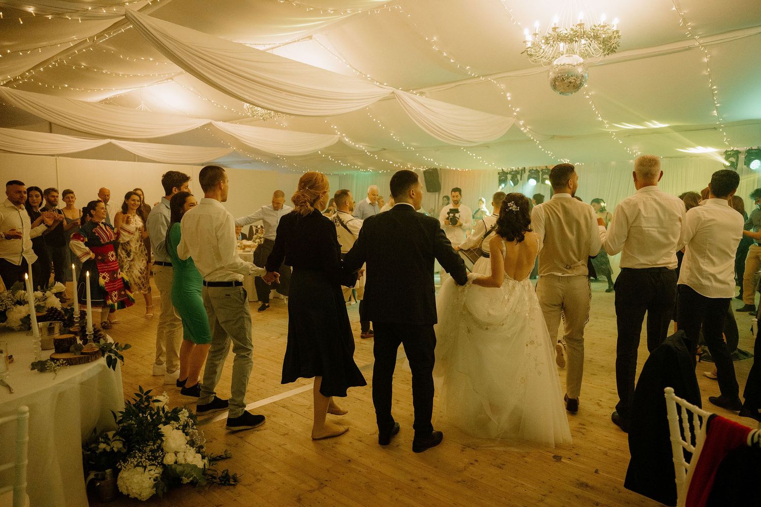 Wedding guests dancing together on a wooden dance floor inside a decorated reception venue with draped ceiling.
