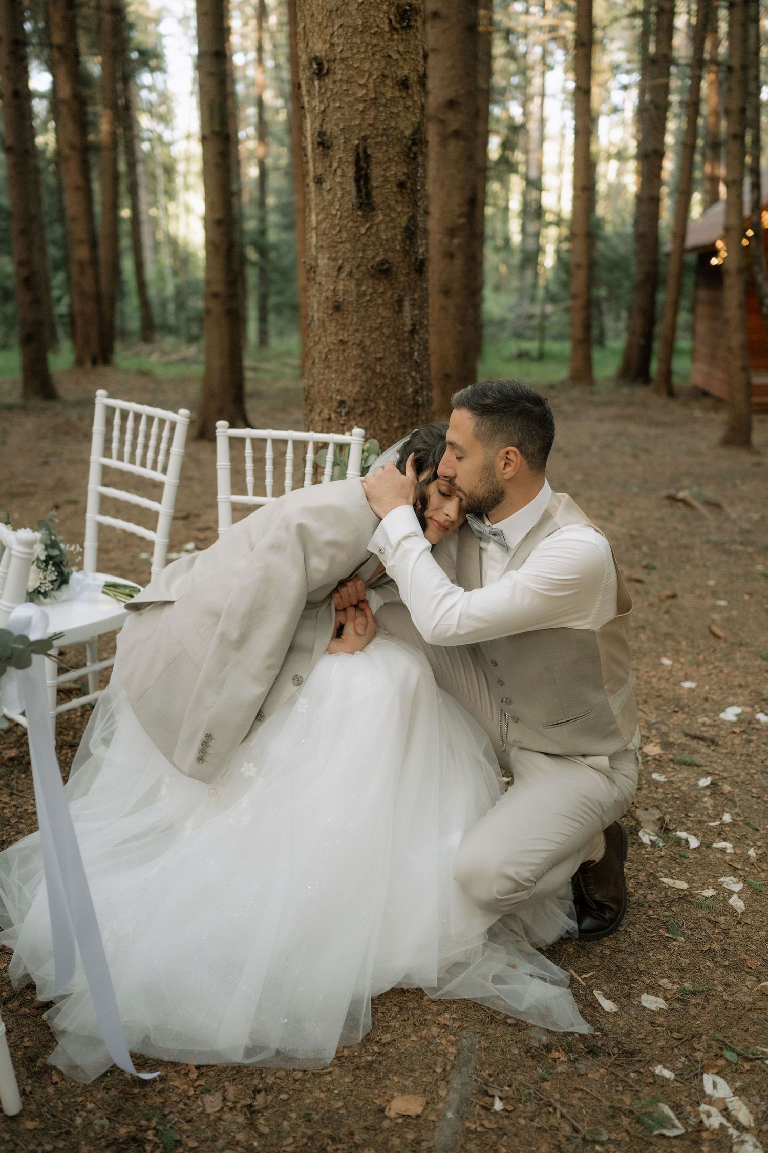 Groom in beige suit tenderly kisses bride in white gown while kneeling in a romantic forest wedding setting.