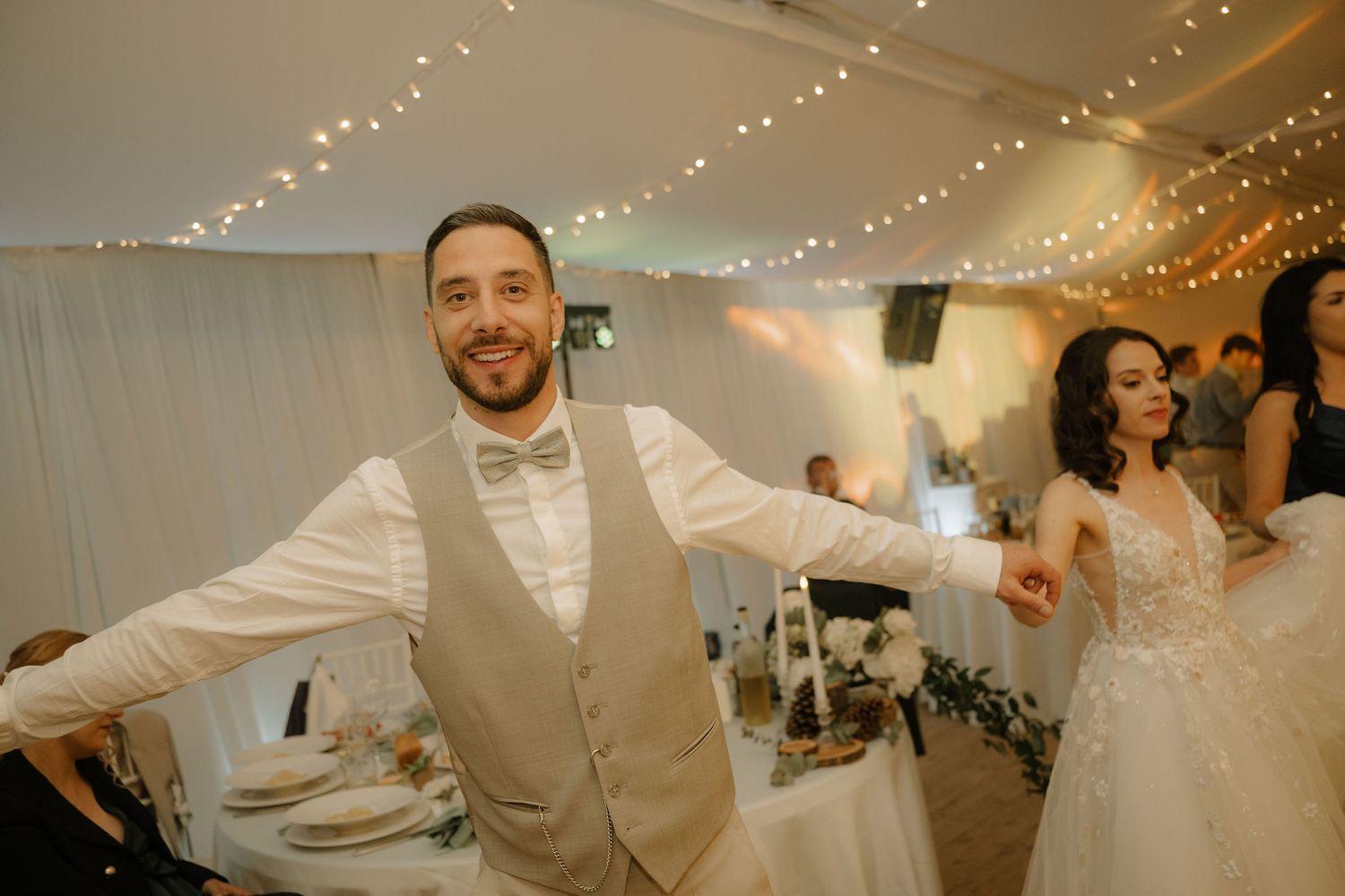 Groom in beige vest and bow tie dancing joyfully at wedding reception with bride in white gown in background.