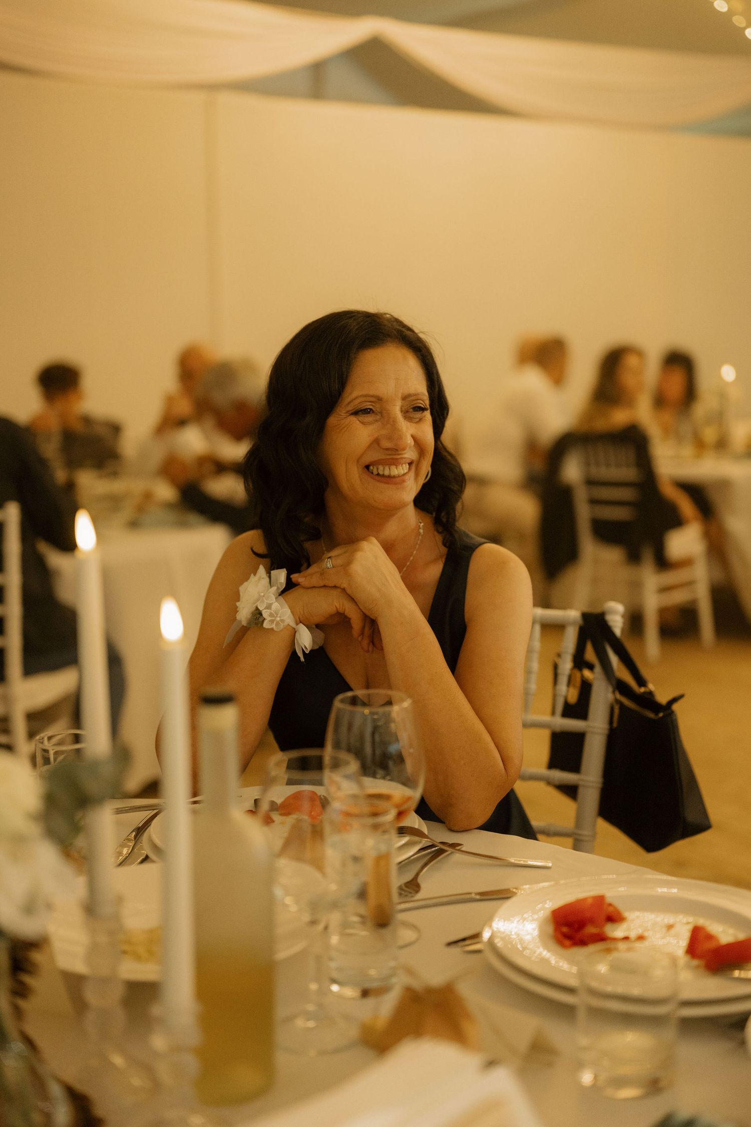 A smiling woman in a black dress sits at an elegantly set dinner table with candles at a formal event.