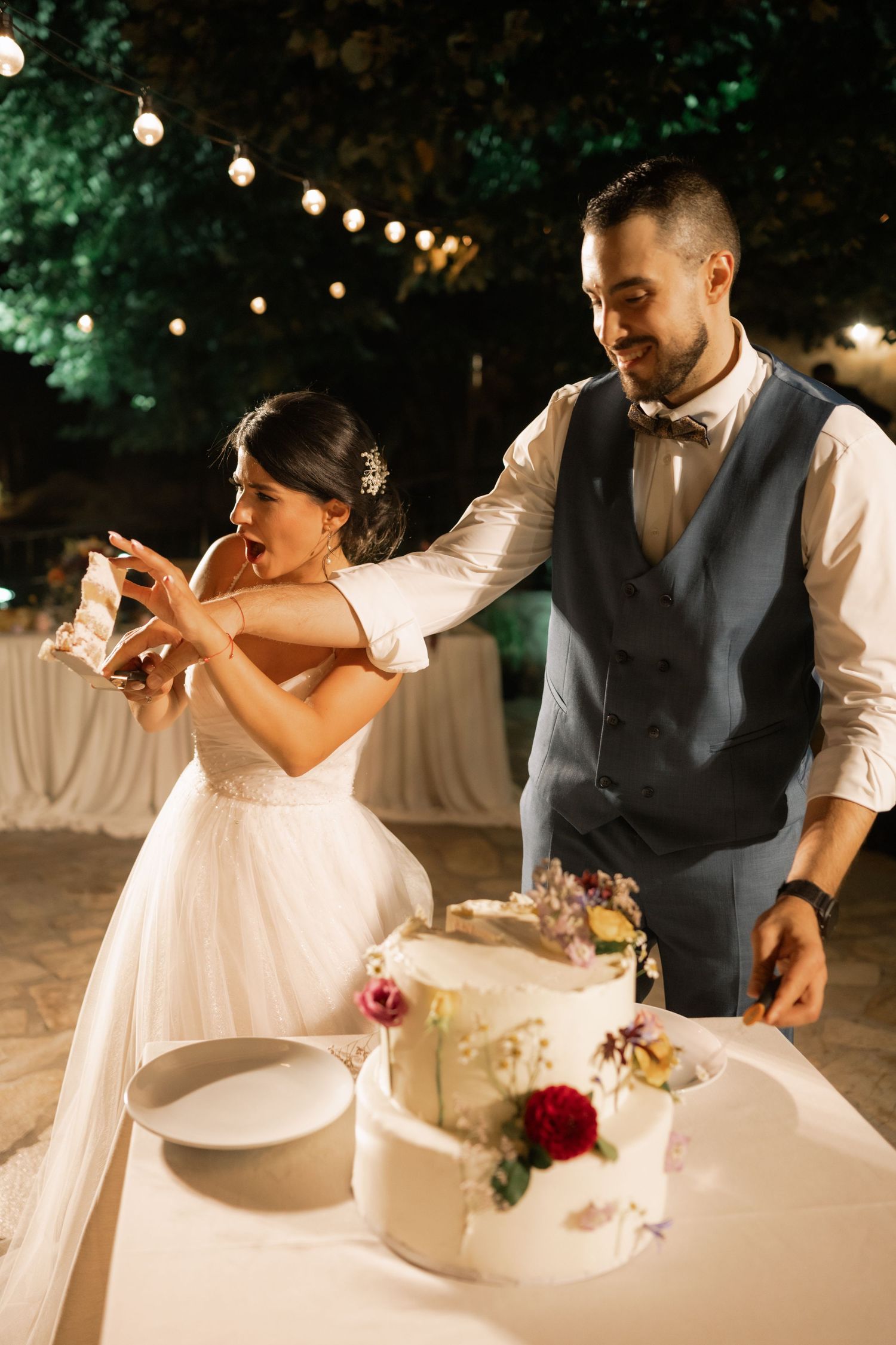 A couple shares a playful moment while cutting their white wedding cake together at an outdoor evening reception.