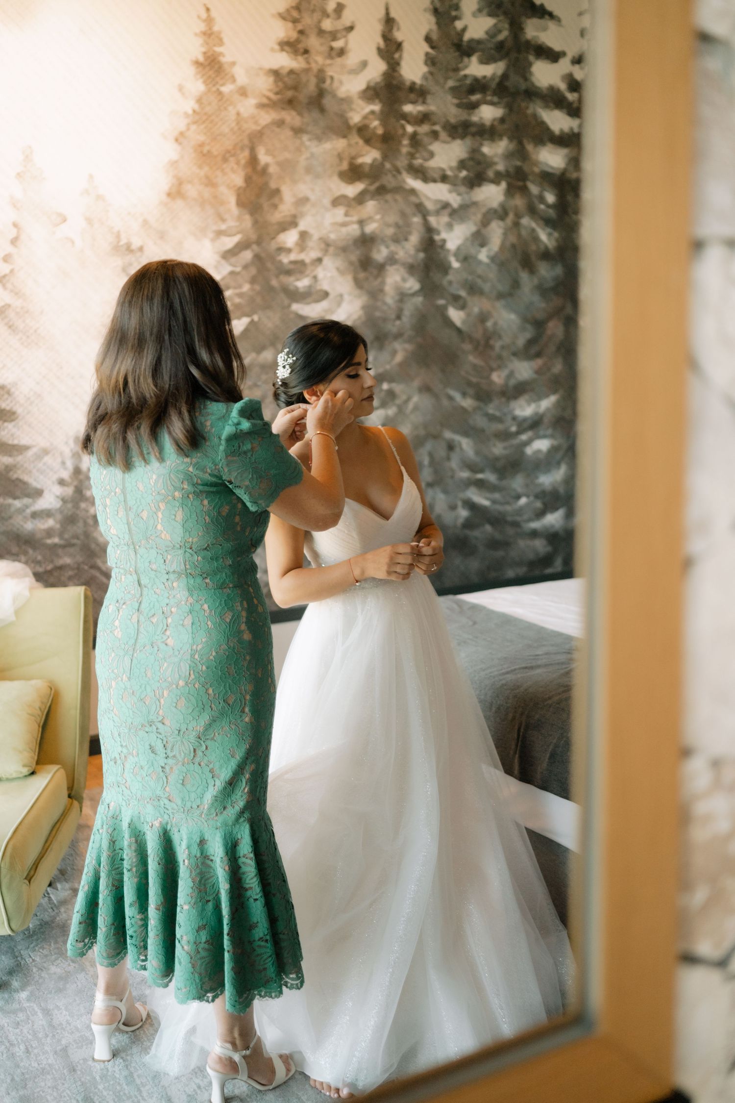 Two people in formal attire, one wearing a mint green lace dress and one in a white gown, look out at snowy evergreens.