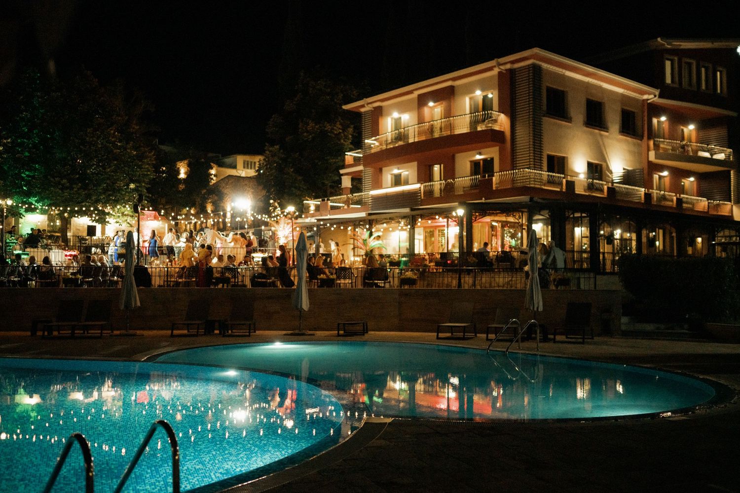 Illuminated resort hotel with outdoor swimming pool reflecting lights at night, surrounded by dining tables and guests.