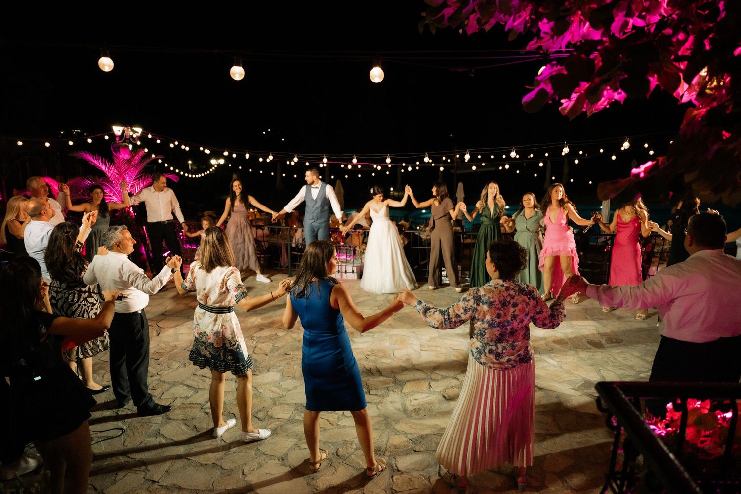 Wedding guests dance together under string lights at an outdoor evening reception with pink floral decorations.