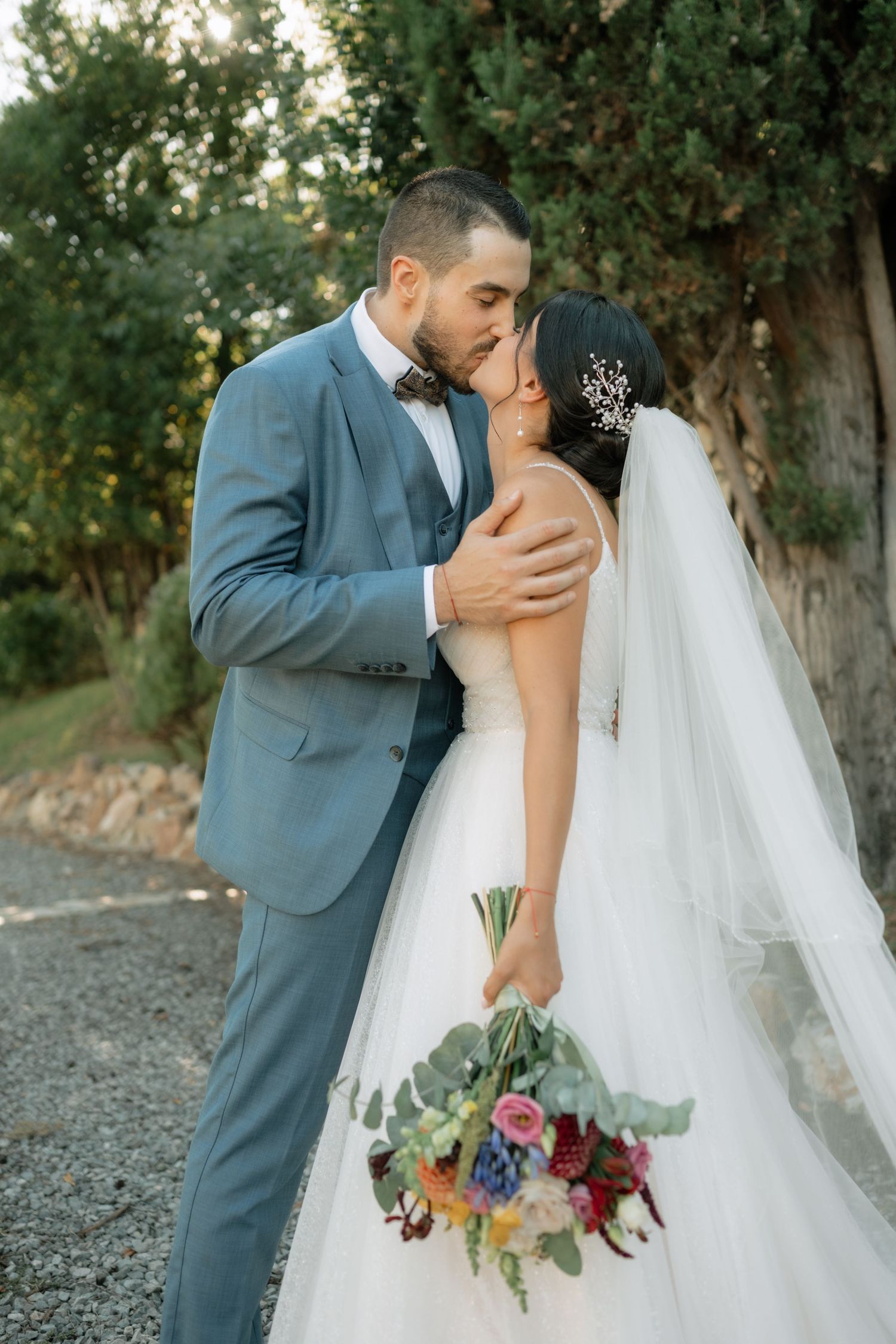 A romantic wedding couple embraces outdoors with the bride holding a vibrant multicolored bouquet against a natural backdrop.